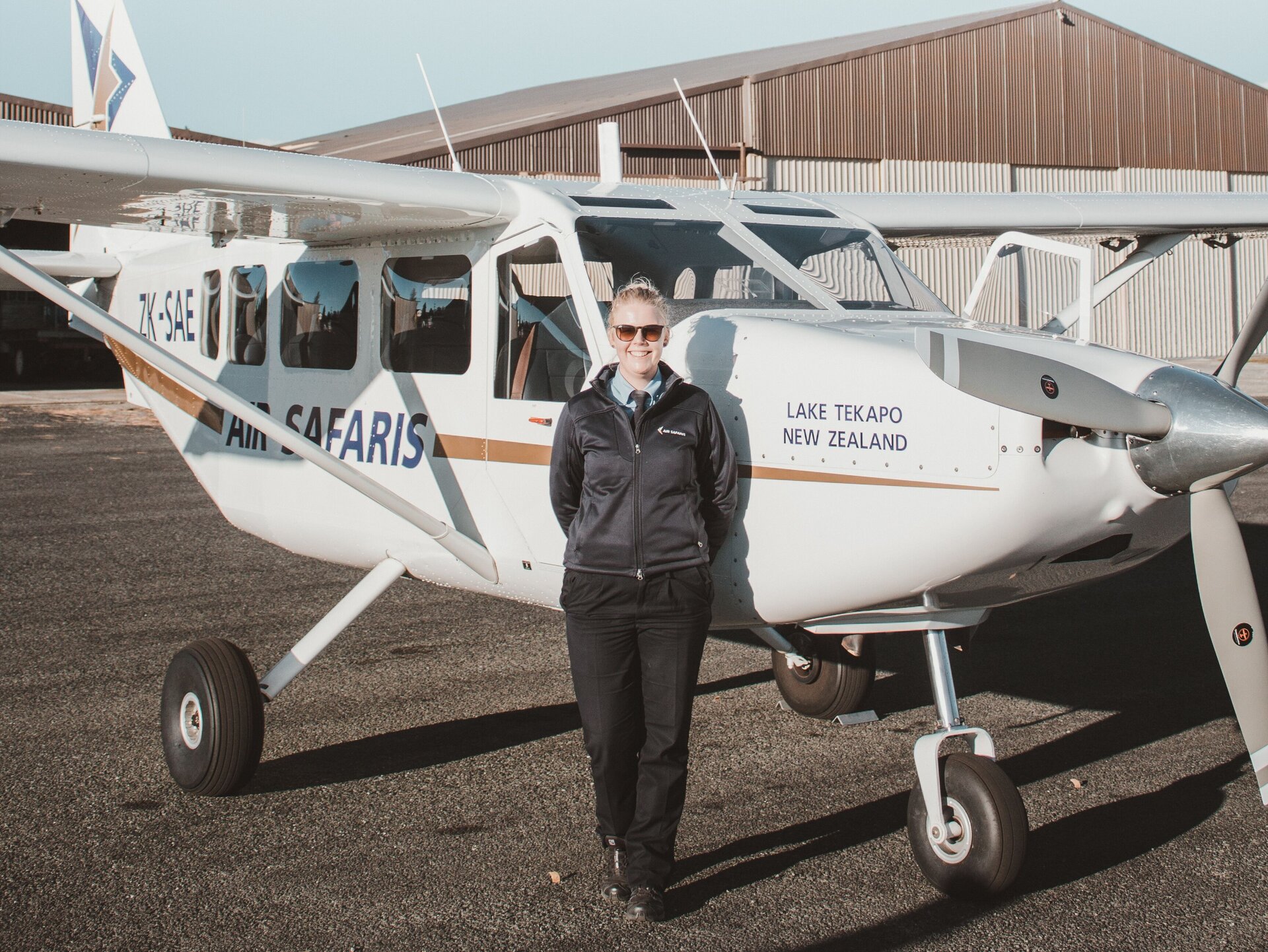 Zdjęcie przedstawia uśmiechniętą, młodą kobietę, stojącą na płycie lotniska i opierającą się o mały samolot. Kobieta ubrana jest w czarną kurtkę, spod której wystaje niebieska koszula i czarny krawat, oraz czarne spodnie. Za nią widoczny jest samolot ze śmigłem na przodzie. Na kadłubie samolotu widoczne są napisy w języku angielskim: AIR SAFARIS, LAKE TEKAPO, NEW ZEALAND. W tle widoczny jest hangar.