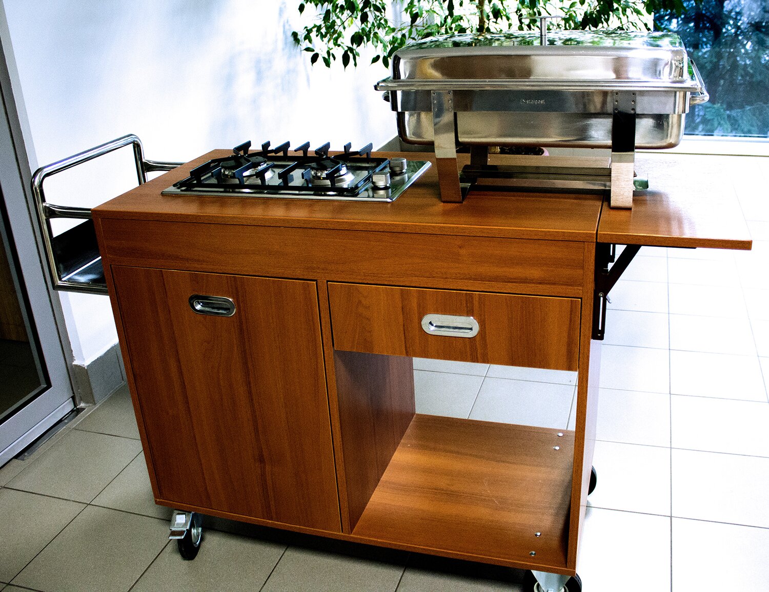 Na zdjęciu znajduje się dystrybutor jezdny grzewczy, przypominający drewniane biurko z szafką po lewej stronie i szufladą po prawej. Blat stołu poszerzony jest o rozkładaną klapkę z prawej strony i metalową półkę z lewej strony. Na blacie znajduje się kuchenka gazowa i podgrzewacz do potraw. Całość konstrukcji na kółkach.