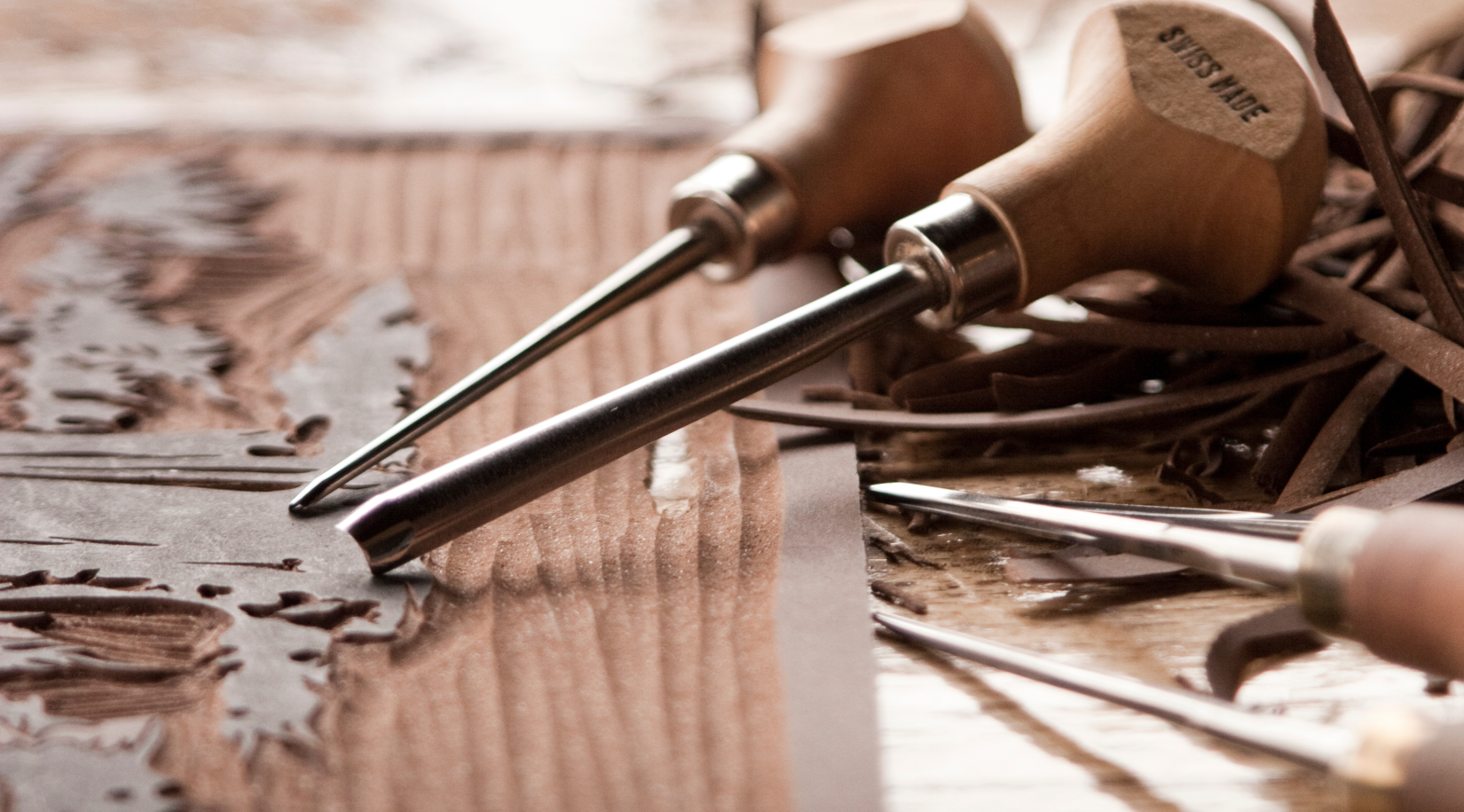 Ilustracja przedstawia dłuta leżące na stole. Pod dłutami znajduje się drewniana deska, na której wyryte są różne wzorki. W tle widać wiórki drewna.