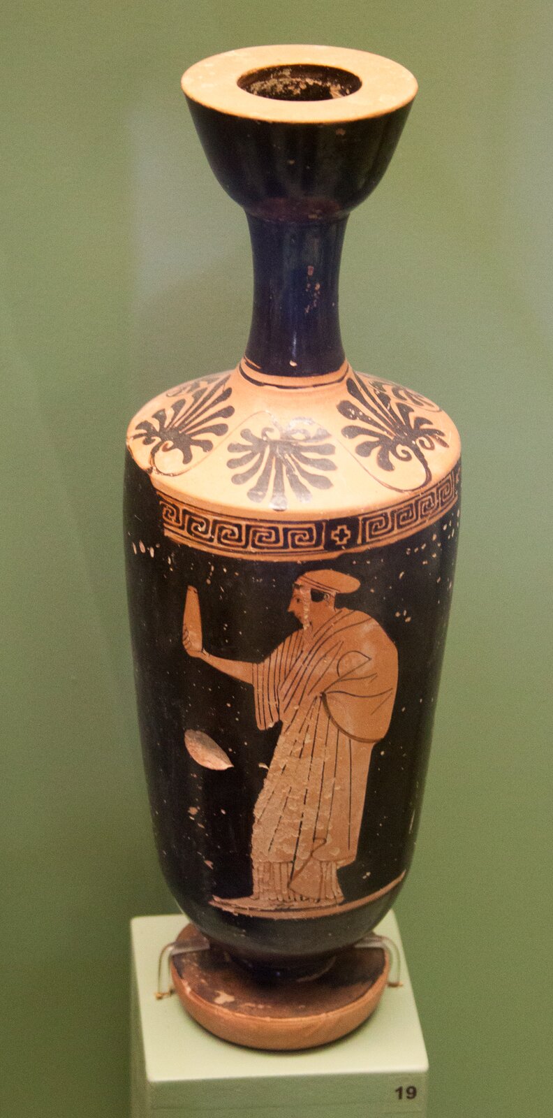 Fotografia przedstawia antyczne naczynie w formie dzbanka, udekorowane malarstwem czerwonofigurowym. Malunek przedstawia mężczyznę ubranego w togę, trzymającego w ręku wysokie naczynie przypominające wazon. W górnej części brzuśca naczynie ozdobione jest wzorem meandrycznym. Nad nim pas z elementem malarstwa czarnofigurowego: motywy roślinne na żółtym tle.