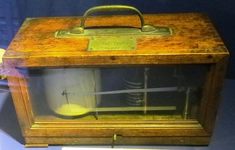 Zdjęcie przedstawia barograf. Urządzenie to znajduje się ono w skrzynce. Posiada wskazówkę, która drgając zapisuje na materiale rolki wychyły.