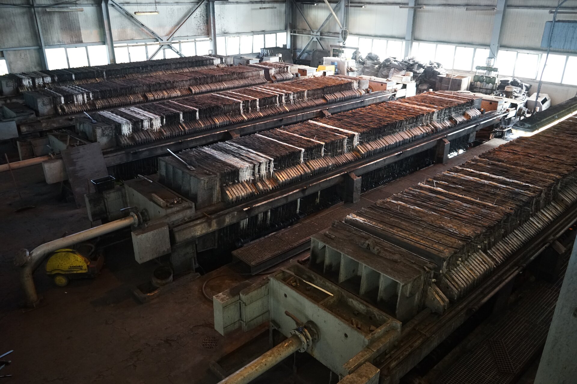 Na zdjęciu widoczne są cztery prasy filtracyjne stojące w hali przemysłowej. Urządzenia widoczne są z góry. Prasa filtracyjna jest to urządzenia filtrujące zagęszczone zawiesiny (szczególnie bardzo drobno uziarnione) na zasadzie przepływu cieczy z filtrowanej zawiesiny przez warstwę porowatą, którą stanowią nagromadzone ziarna fazy stałej, a przepływ ten wywołany jest różnicą ciśnień przed i za przegrodą filtracyjną.Podstawą urządzenia jest podłużna prostopadłościenna skrzynia. W środku skrzyni znajdują się filtry. Filtry są o kształcie cienkich prostopadłościanów. Z przodu urządzenia widoczna jest rura. Pomiędzy urządzeniami utworzony jest przejście.