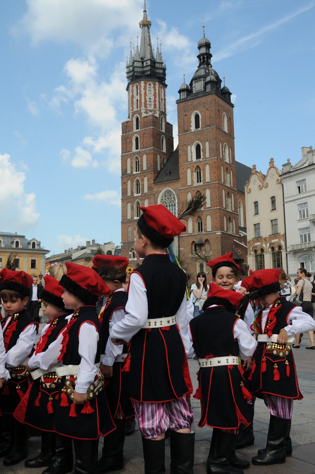 Zdjęcie przedstawia grupę przedszkolaków, którzy są ubrani w tradycyjne krakowskie stroje. W tle widać Kościół Mariacki. 