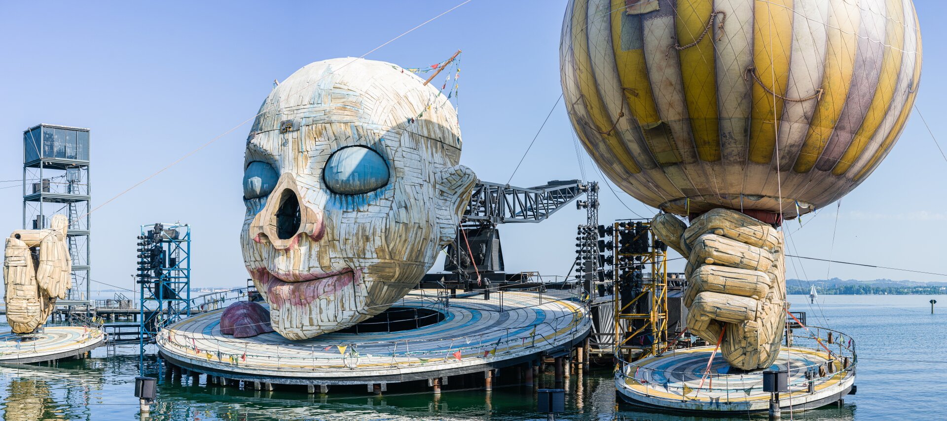 Ilustracja przedstawia scenografię do opery „Rigoletto”, autorstwa Giuseppe Verdi. Na pierwszym planie, na środku fotografii znajduje się wielka głowa. Po jej bokach są 2 dłonie. Na twarzy znajdują się, pomalowane niebieską farbą, zamknięte oczy oraz pomalowane na czerwono, uśmiechnięte usta. W miejscu gdzie powinien być nos znajduje się dziura. Czerwony nos leży po lewej stronie, przy podbródku. Po dwóch stronach głowy wystaje belka. Przyczepione są do niej girlandy w kolorowe trójkąty, których drugi koniec przyczepiony jest do głowy. Poniżej  głowy znajduje się okrągła platforma z barierką, do której przyczepione są girlandy z kolorowymi trójkątami. Platforma jest w kolorach: surowego jasnego drewna, żółtym, niebieskim i szarym. Po prawej stronie ilustracji znajduje się duża dłoń trzymająca balon. Dłoń jest w kolorze jasnego drewna, a balon jest kremowo‑żółty w pionowe pasy. Z lewej strony ilustracji znajduje się druga duża dłoń, której palce są zamknięte. Pod każdą dłonią jest okrągła platforma. Są one podobne do tej, która jest pod głową, ale mają mniejszą średnicę. Na drugim planie znajdują się sprzęty oświetleniowe na wysokich stelażach. Po lewej stronie fotografii, między dłonią a głową, jest niebieski stelaż. Po prawej stronie fotografii, między dłonią z balonem a głową, jest żółty stelaż. Za głową znajduje się czarny stelaż. Po lewej stronie fotografii, za dłonią jest 5‑cio poziomowy stelaż z drabinami. Ostatni poziom jest przeszklony. Scenografia znajduje się na obiekcie wodnym. W tle jest błękitne niebo, a w oddali drzewa.