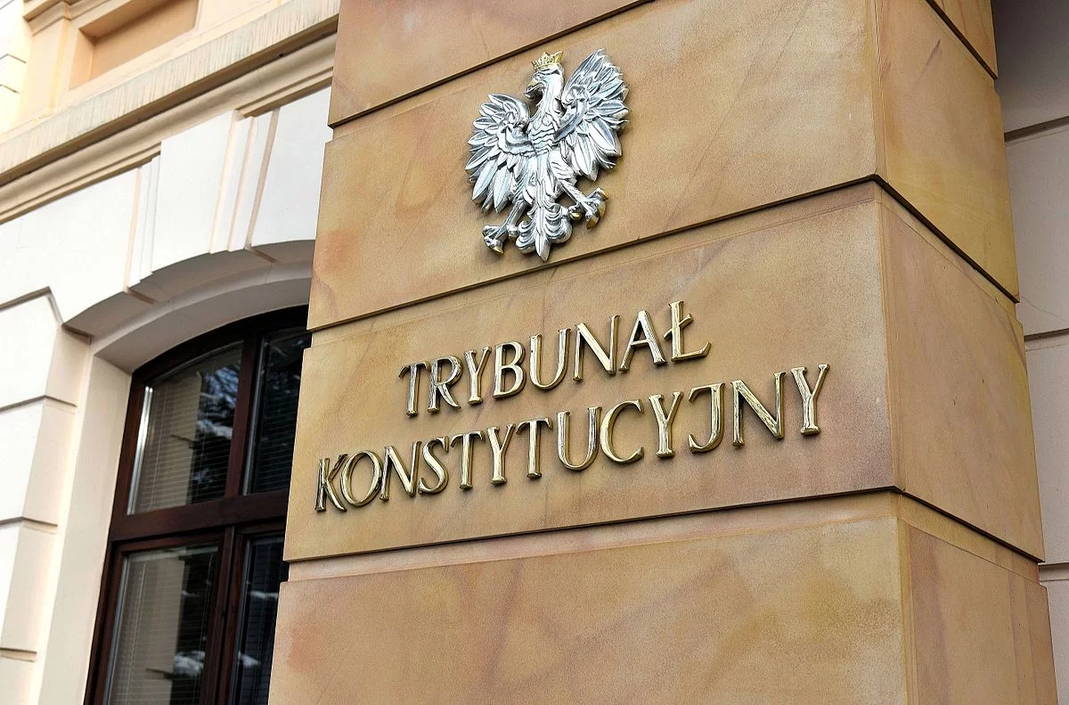 Na zdjęciu znajduje się masywna ściana z napisem Trybunał Konstytucyjny oraz srebrnym godłem Polski. Po lewej widoczne jest drewniane okno.