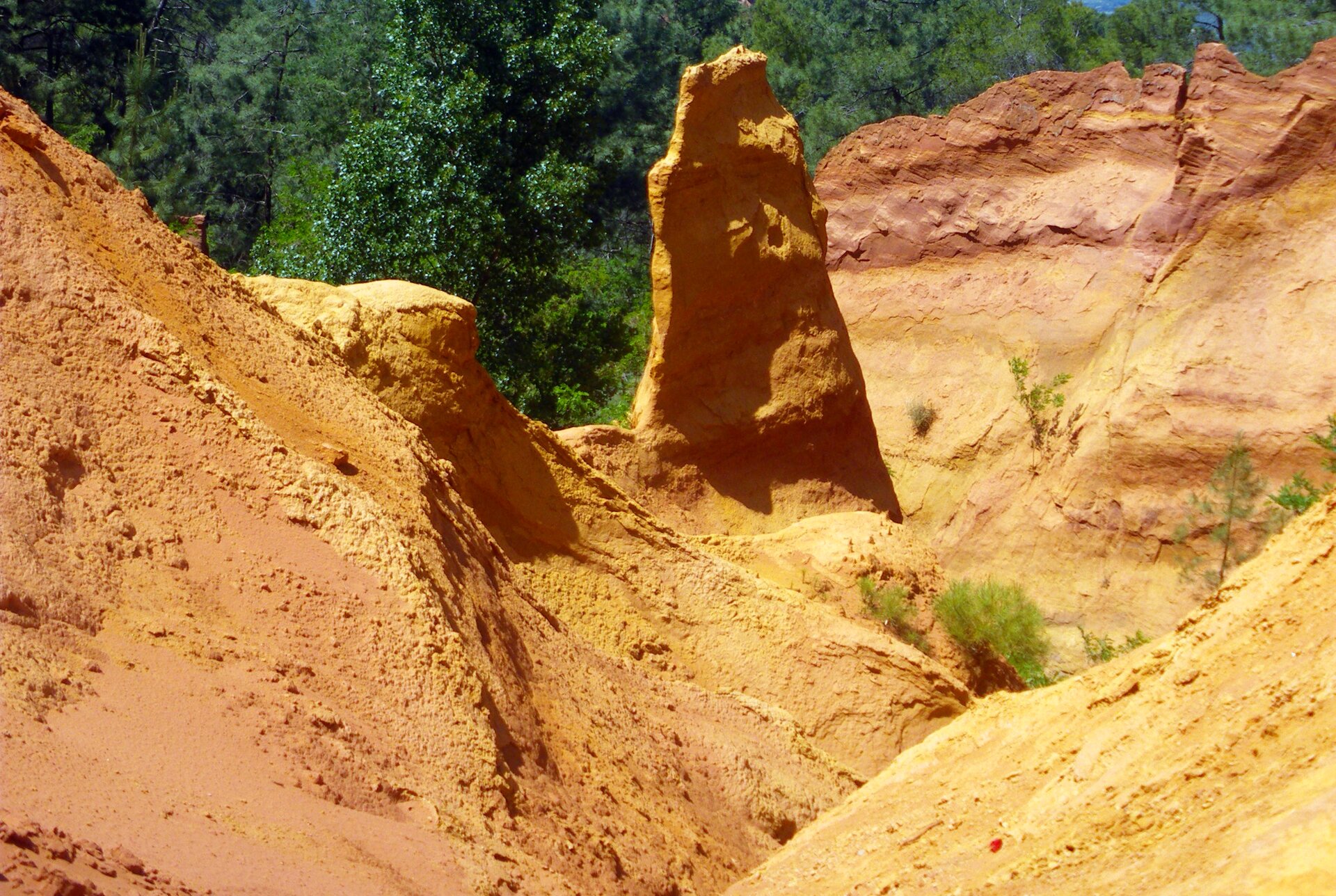 Kliknij, aby powiększyć. Fotografia przedstawia skały ochrowe. Pomarańczowe skały w różnych odcieniach. W centrum fotografii wystająca skała. W tle zielony las mieszany.