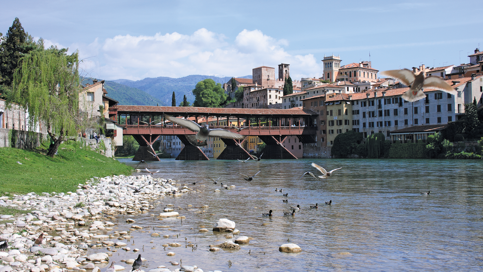 Zdjęcie przedstawia widok na miasteczko Bassano del Grappa w Wenecji Euganejskiej. Na pierwszym planie widoczna jest rzeka. W oddali znajduje się stary most oraz budynki starego miasta.