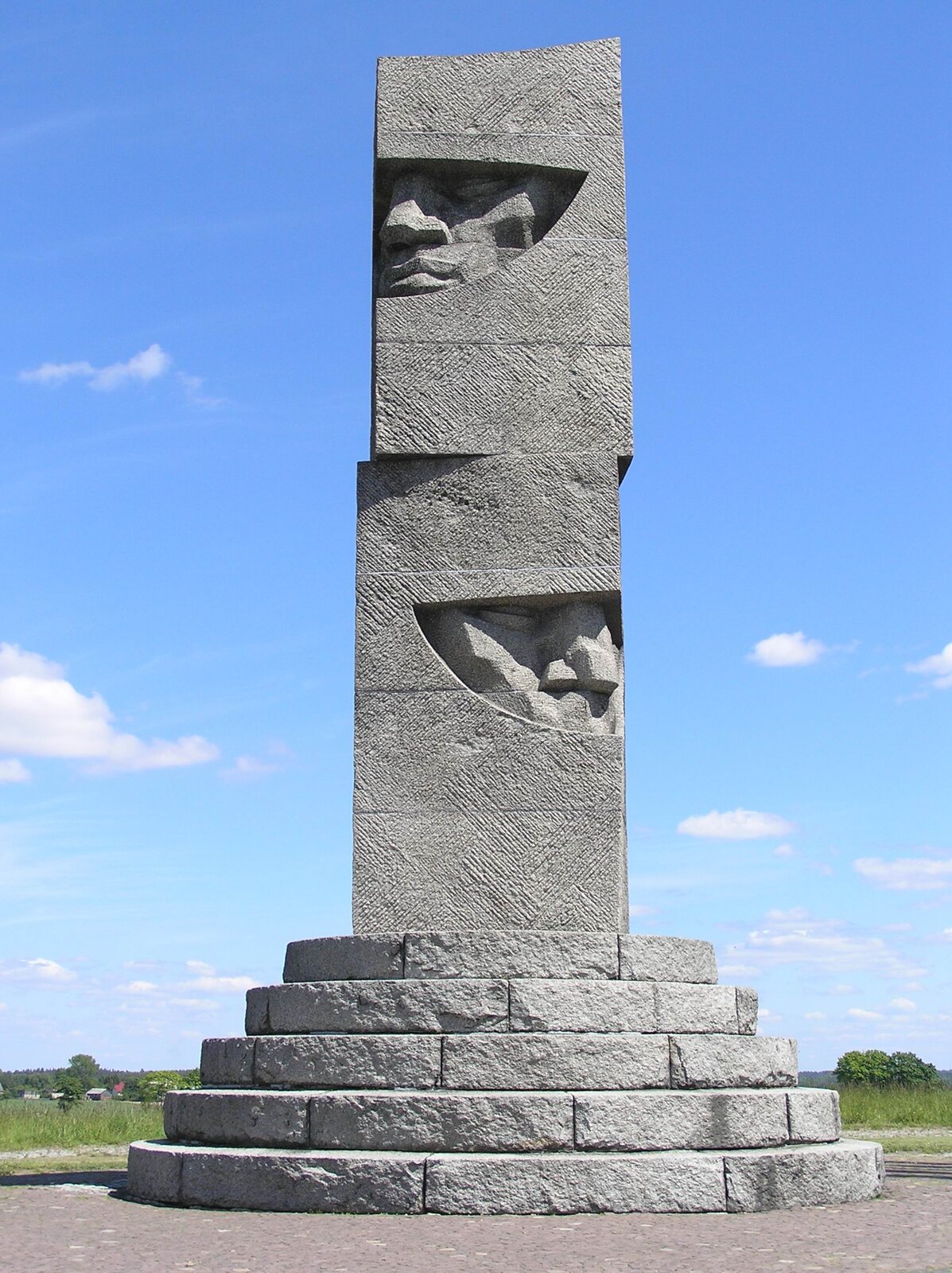 Na zdjęciu widać pomnik upamiętniający bitwę pod Grunwaldem. Jest on kamienny, stoi na cokole w formie pięciu okrągłych schodów. Pomnik ma kształt wysokiego prostopadłościanu, na którego krawędziach widać fragmenty dwóch twarzy - policzki, oczy, usta i nosy. W głębi widać pas  roślinności i niebieskie niebo.