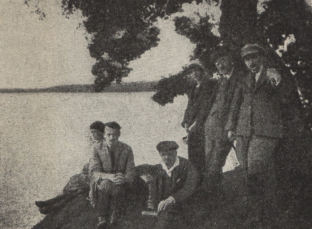 Kliknij, aby powiększyć. Czarno‑biała fotografia, widok jeziora, na pierwszym planie znajduje się grupa osób, trzy siedzą, trzech mężczyzn stoi, jeden z nich opiera się o drzewo. Ubrani są w koszule, marynarki i materiałowe spodnie. Dwóch stojących mężczyzn i jeden siedzący mają na głowach kaszkiety.