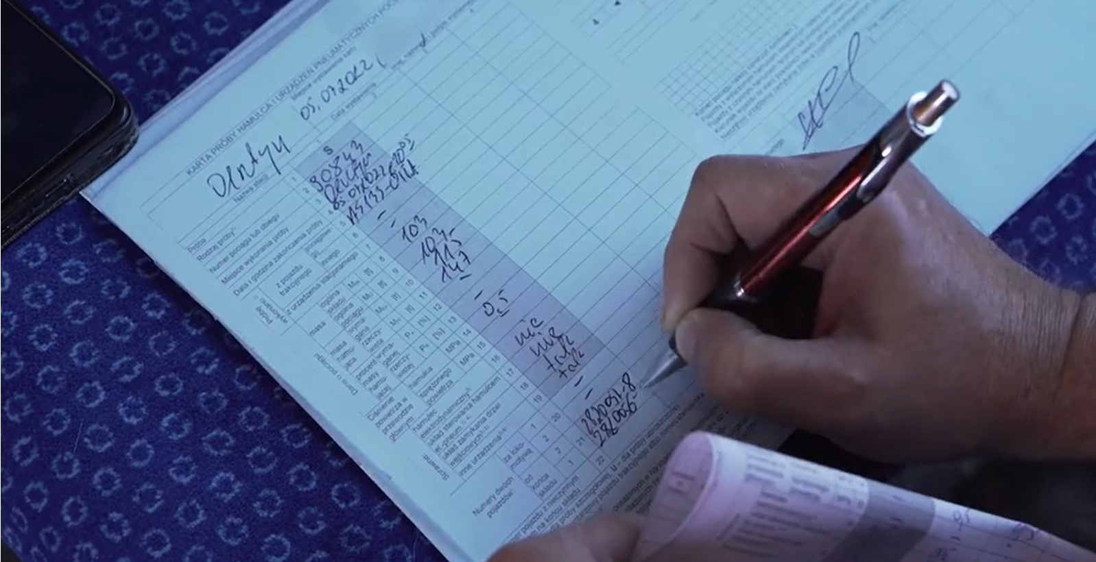 Zdjęcie przedstawia wypełnianą kartę próby hamulca. Widoczny jest dokument, w którego rubrykach ręcznie wpisywane są dane zebrane w czasie próby hamulca. Widać rękę trzymającą długopis, która wpisuje dane w rubrykach kolumny dokumentu.