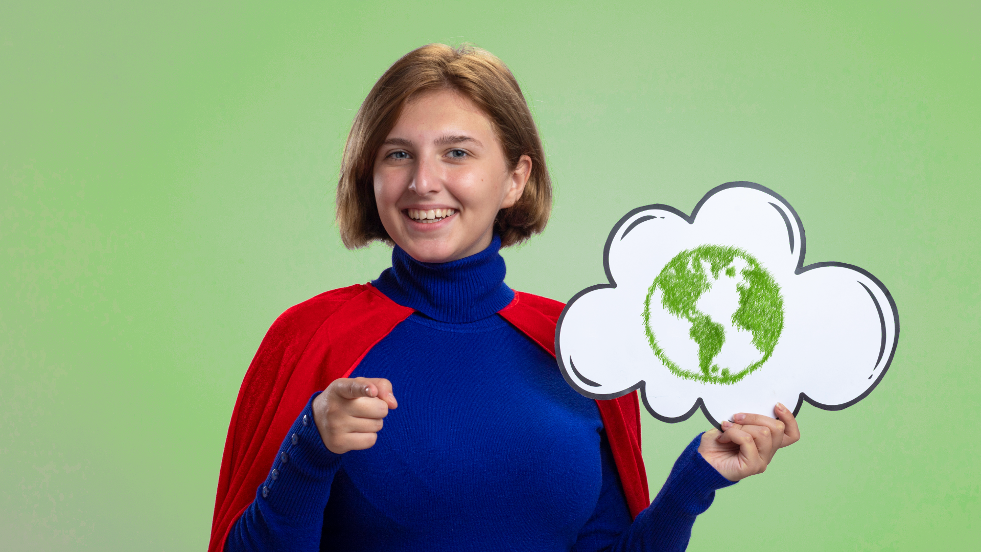 Zdjęcie przedstawia uśmiechniętą kobietę w stroju superbohaterki. Kobieta ma na sobie golf oraz pelerynę. Palec wskazujący jednej dłoni kieruje w stronę kadru. W drugiej dłoni trzyma dymek, w którym znajduje się kula ziemska.