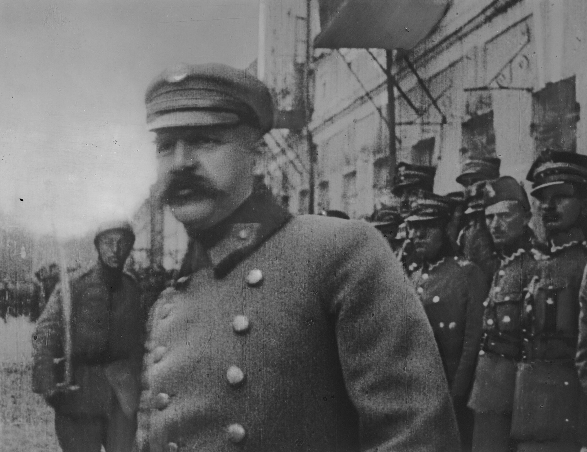 Zdjęcie przedstawia Józefa Piłsudskiego. Znajduje się on na pierwszym planie. Ma mundur oraz czapkę z daszkiem. Za nim stoją żołnierze w podobnych ubraniach. Stoją pod ścianą budynku.