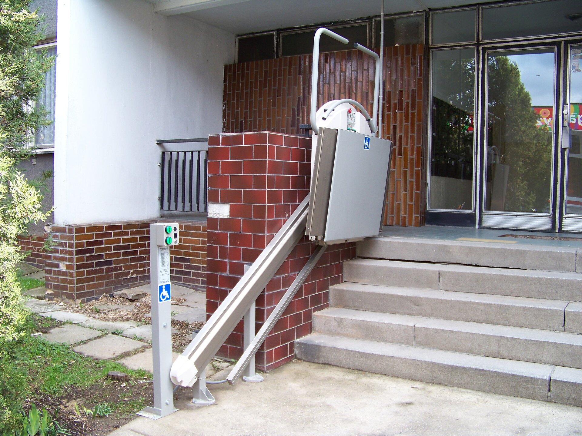 Na zdjęciu widoczne wejście do budynku. Przy ścianie bocznej schodów, przymocowane dwie szyny wzdłuż których porusza się zawieszona platforma do transportu dla niepełnosprawnych. Z boku widoczny panel z przyciskami sterowania oraz instrukcja użytkowania.