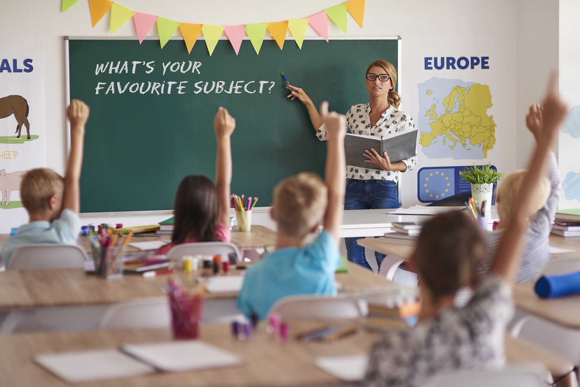 Zdjęcie przedstawia uczniów w trakcie lekcji. W tle widoczna jest kobieta stojąca tyłem do tablicy, trzymająca w lewej ręce otwartą dużą książkę, a w prawej ręce długopis, którym wskazuje na napis znajdujący się na tablicy: WHAT'S YOUR FAVOURITE SUBJECT?. Po prawej stronie tablicy, na ścianie, widoczna jest mapa Europy, nad którą znajduje się napis w języku angielskim: EUROPE. Na pierwszym planie widać szkolne ławki, na których leżą różne przybory do malowania. Przy ławkach siedzą dzieci, wszystkie mają uniesioną prawą rękę.