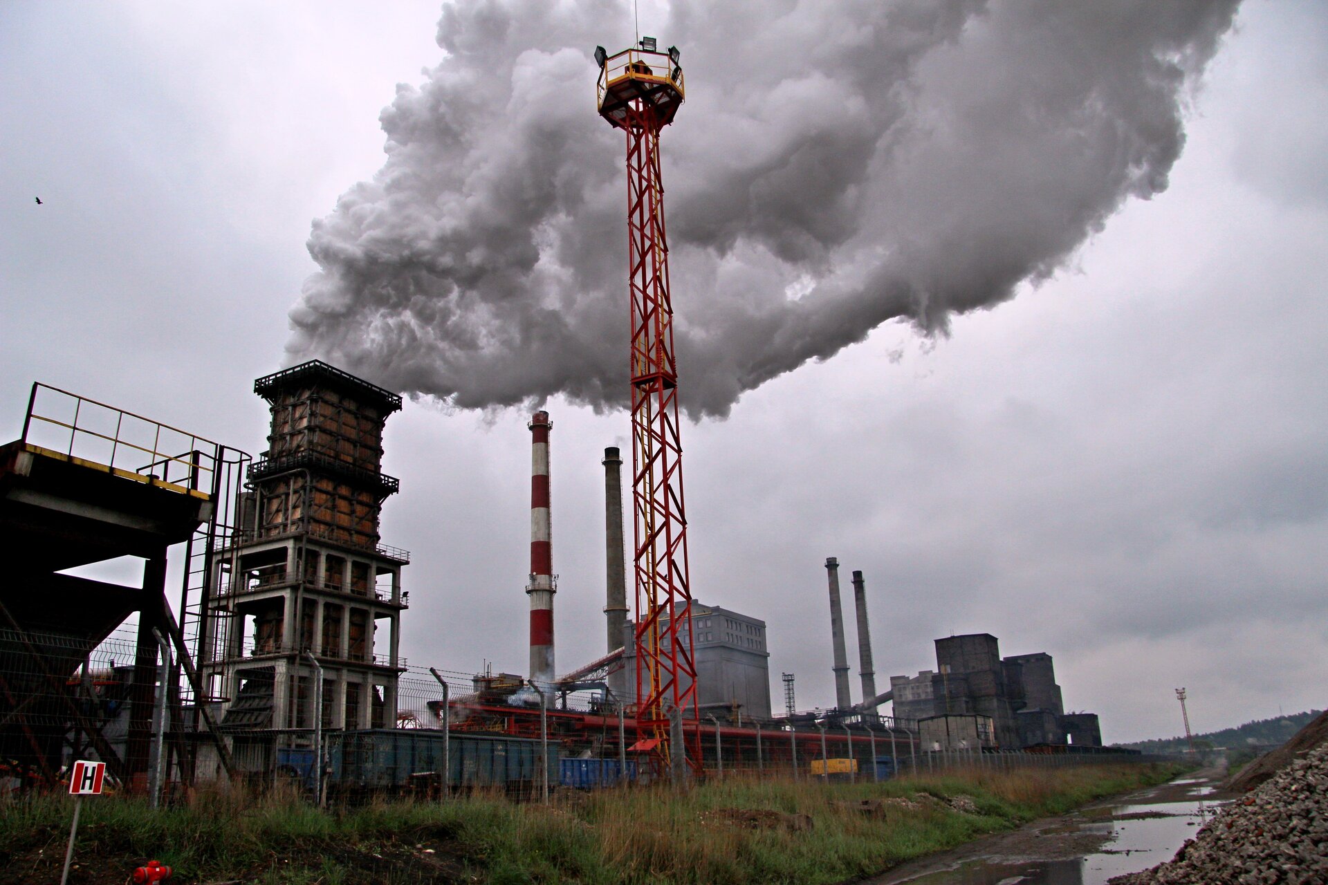 Zdjęcie przedstawia widok z oddali na dużą fabrykę z licznymi wysokimi kominami. Z niektórych kominów wydobywa się biały dym.