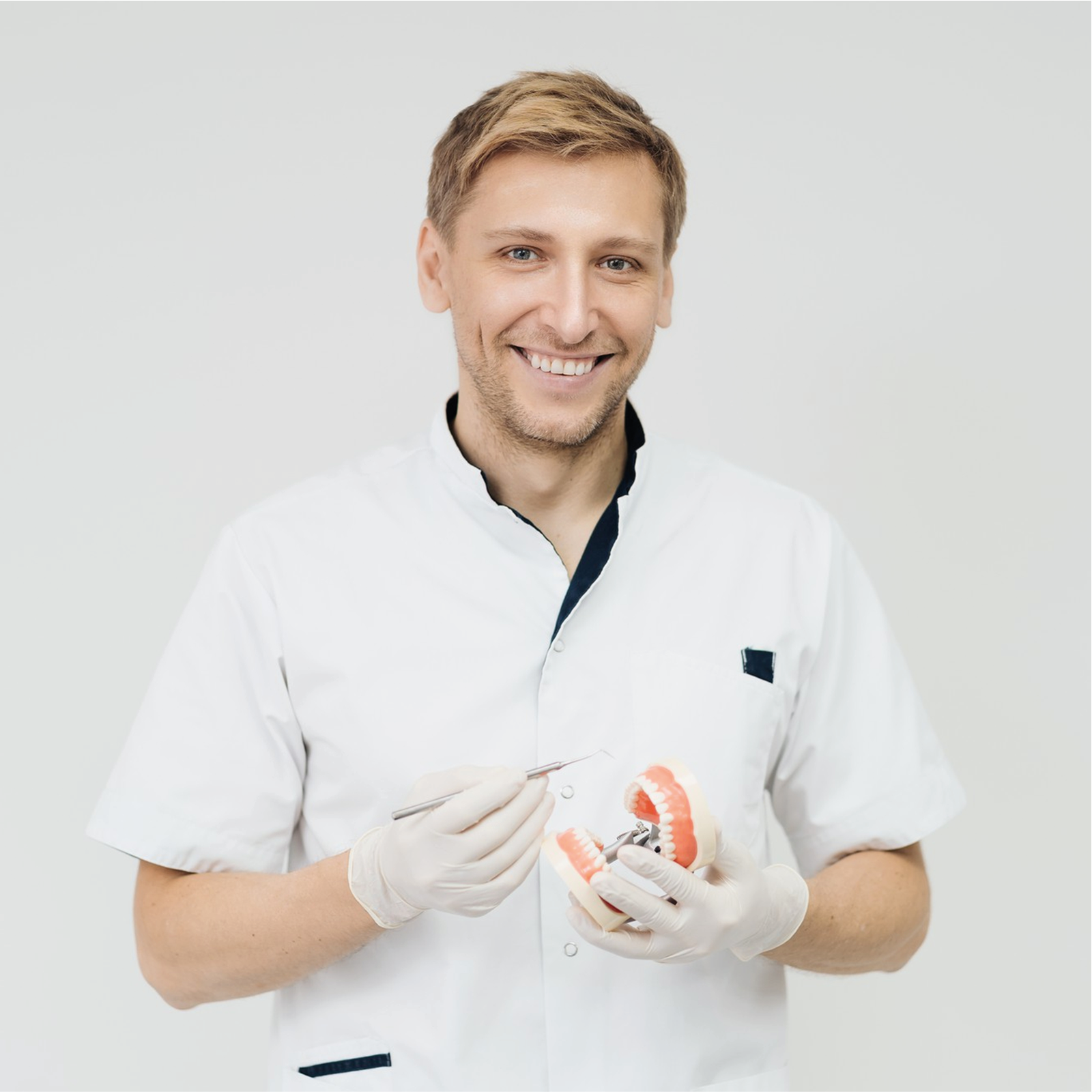 Zdjęcie przedstawia mężczyznę w białej, zapinanej koszuli i rękawiczkach, który trzyma sztuczną szczękę i narzędzie do badania zębów.
