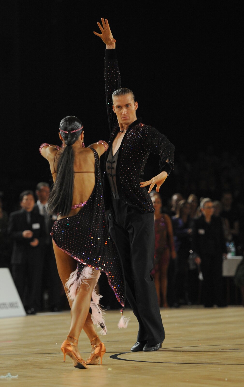 Fotografia przedstawia parę w tańcu. Kobieta ma na sobie czarną sukienkę z kolorowymi, błyszczącymi elementami. Sukienka wykończona jest białymi frędzlami. Na stopach ma złote sandały na obcasie. Ma duże, różowe kolczyki, a na obu ramionach ma błyszczące bransoletki w kolorze różowym. Kobieta ma czarne włosy zaczesane do tyłu w kucyk oraz różową, błyszczącą opaskę. Stoi tyłem i ma skrzyżowane nogi. Po prawej stronie stroi mężczyzna. Ma czarną marynarkę z kolorowymi, błyszczącymi elementami, czarną, przeźroczystą bluzkę, czarne spodnie oraz zakryte buty. Jego lewa ręka jest na wysokości talii, a prawa podniesiona do góry. Para znajduje się na jasnym parkiecie z czarnymi okręgami. W tle znajduje się widownia. Tło jest rozmyte, niewyraźne. 