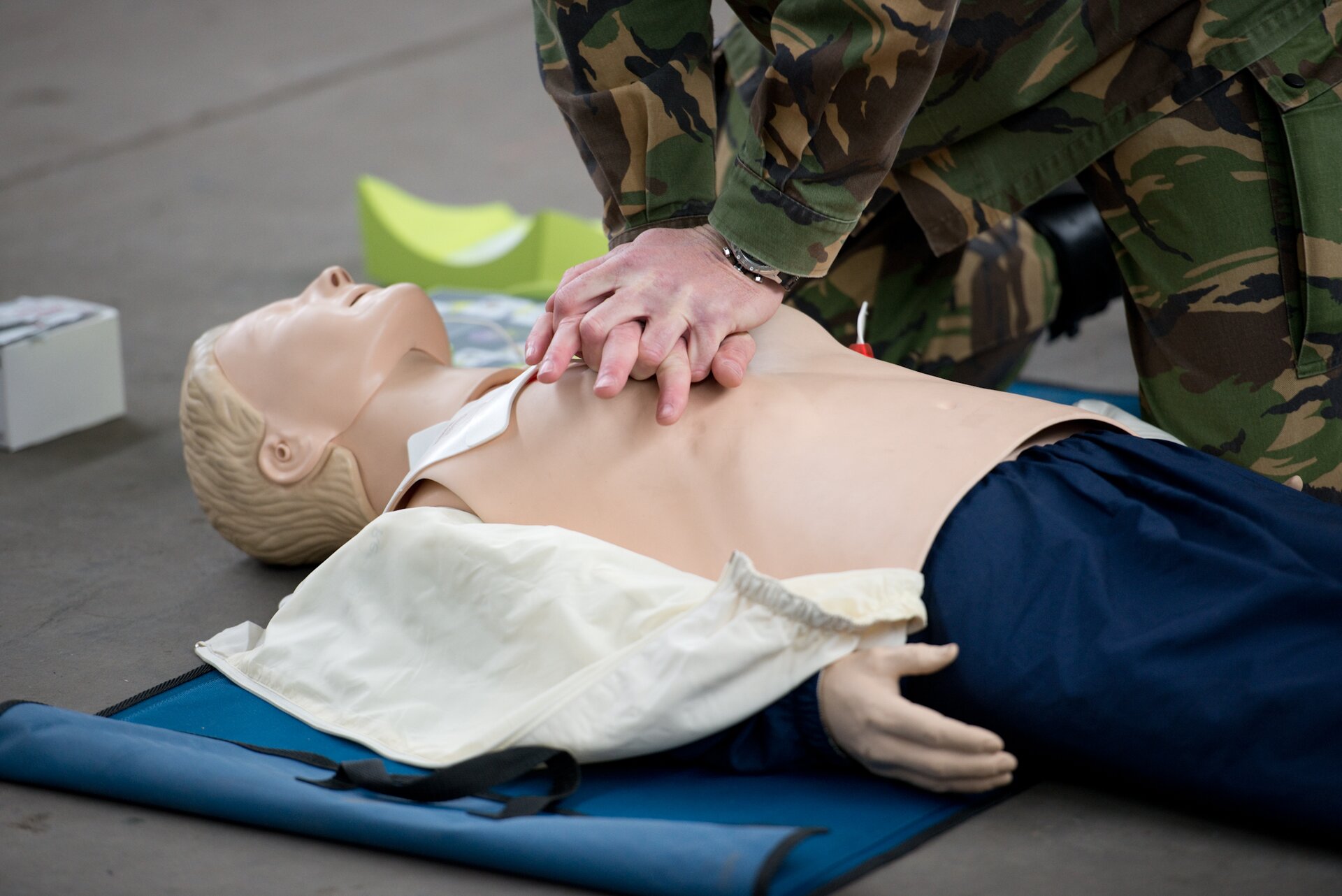 Kliknij, aby powiększyć. Zdjęcie przedstawia fantom męski, nad którym mężczyzna ubrany w mundur moro pokazuje reanimację. Fantom ubrany jest w granatowe spodnie i białą koszulę, która została rozpięta. Leży na niebieskiej, prostokątnej macie. Mężczyzna w mundurze klęka pochylając się nad fantomem. Dłonie trzyma splecione na jego klatce piersiowej. Tło jest rozmazane.