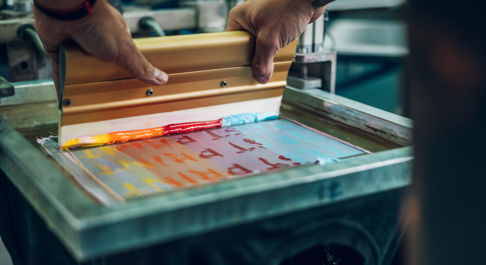 Ilustracja przedstawia maszynę drukarską. Na zdjęciu znajdują się ręce trzymające maszynę do druku. Na maszynie znajdują się różnokolorowe napisy.