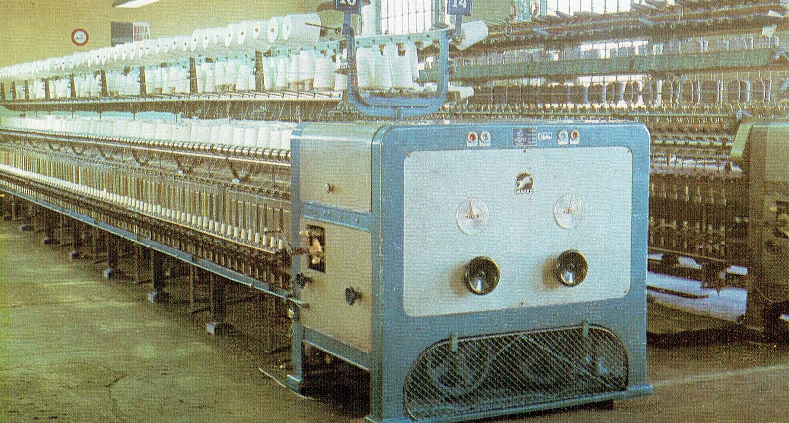 Zdjęcie przedstawia skręcarkę. Na pierwszym planie widoczny panel sterujący maszyny oraz silnik w obudowie. Na drugim planie długa maszyna z ustawionymi obok siebie szpulkami nici w trzech rzędach. W tle druga skręcarka. 