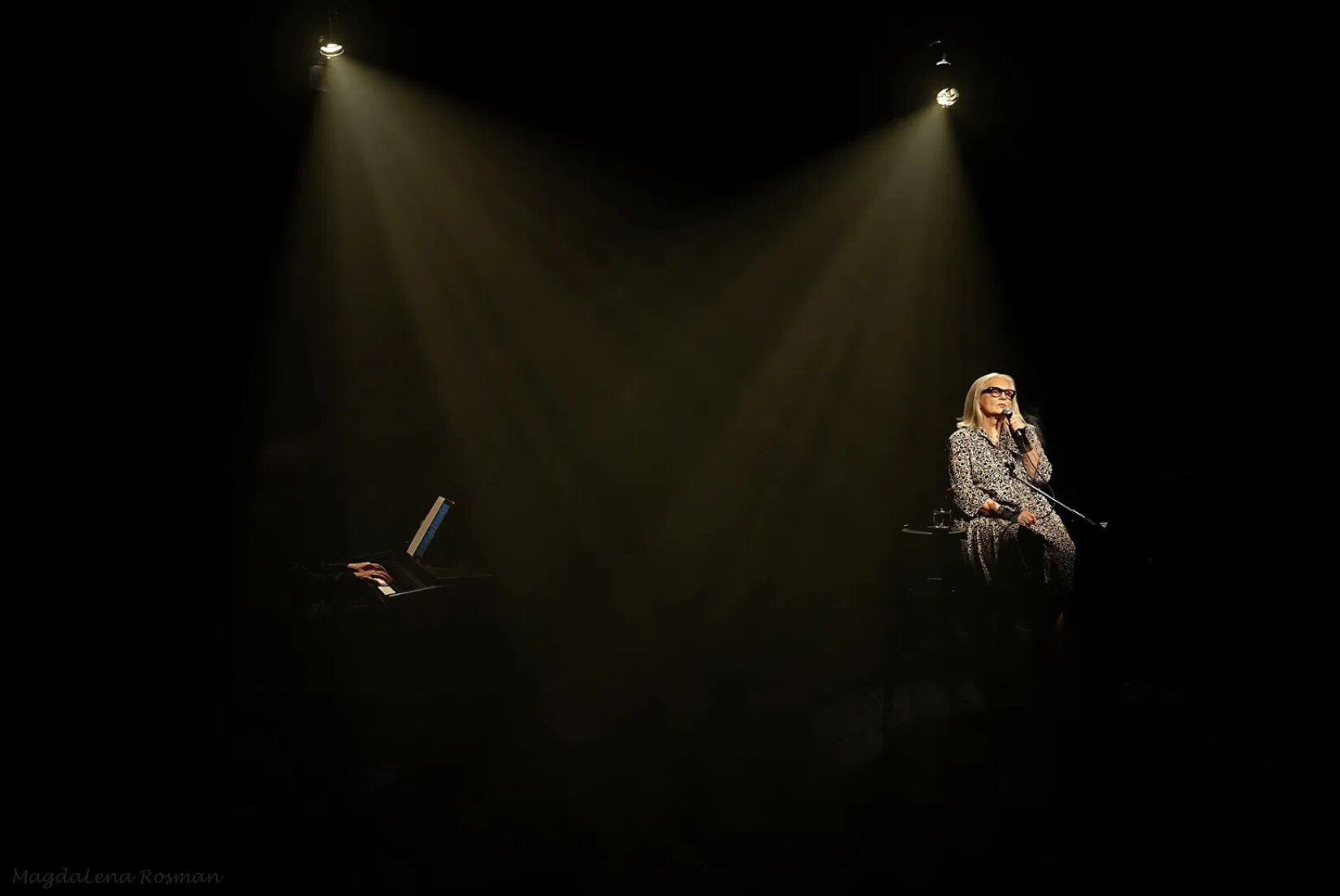 Zdjęcie przedstawia muzyka grającego na instrumencie klawiszowym oraz blondwłosą kobietę w średnim wieku siedzącą na krześle. Ubrana jest w kombinezon w biało‑czarne wzory geometryczne. W lewej dłoni trzyma mikrofon a w prawej przewód mikrofonu. Fotografia jest ciemna, od góry scenę rozświetlają dwie lampy, które dają jasno brązową poświatę. 