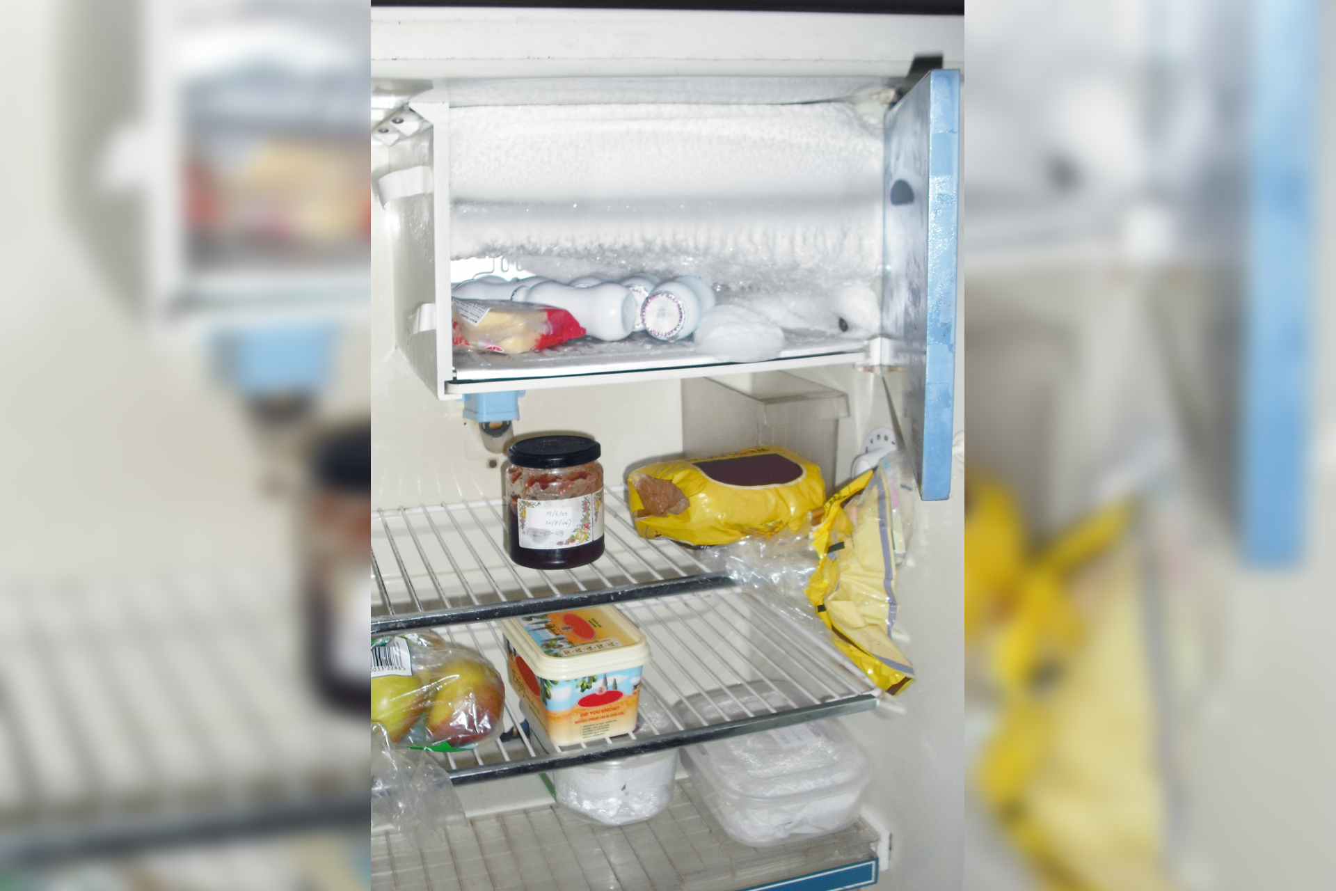 Zdjęcie ukazujące otwartą, dużą lodówkę. W lodówce na półkach znajduje się niewiele produktów spożywczych: chleb, dżem, jabłka, pojemniki plastikowe z żywnością. Zamrażalnik bardzo oblodzony z kilkoma produktami.