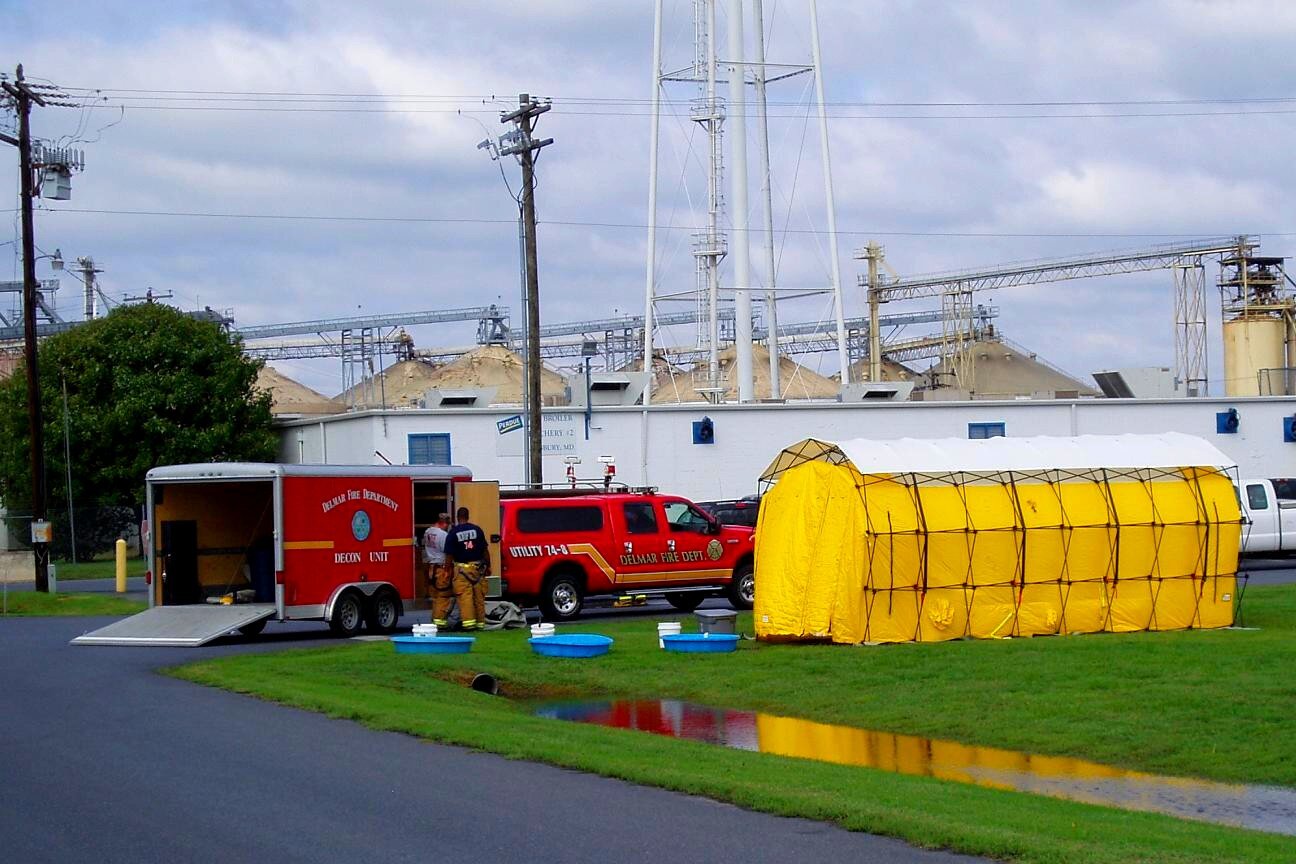 Zdjęcie przedstawia wóz strażacki z przyczepą. Obok niego znajdują się strażacy oraz żółty namiot.