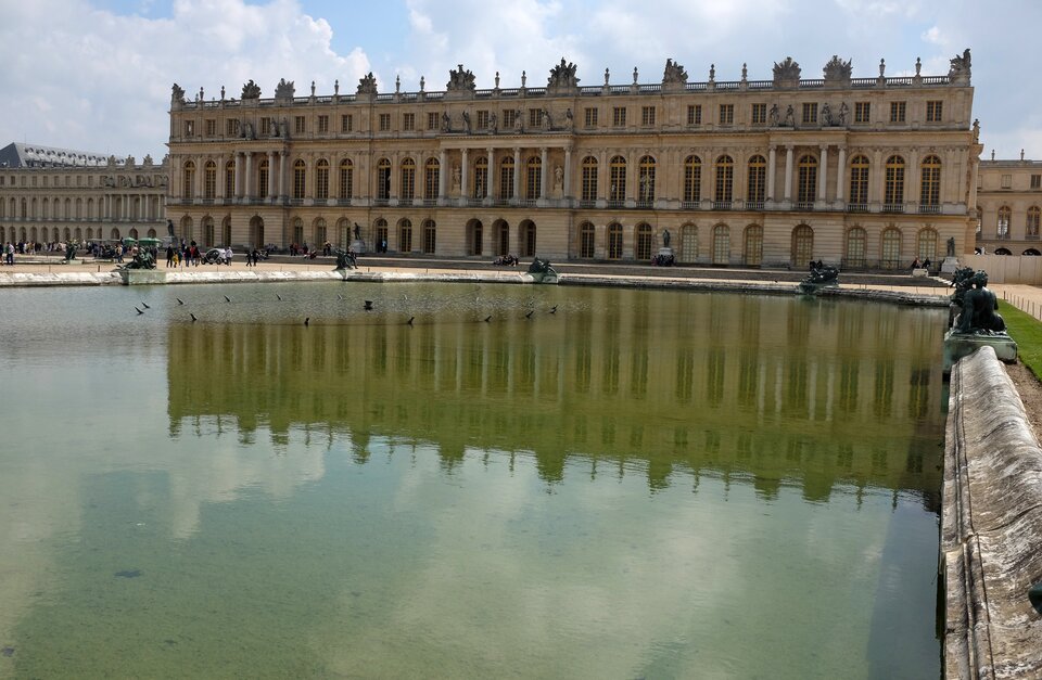 Na zdjęciu znajduje się fragment pałacu w Wersalu. Budynek ma kształt prostokąta, składa się z dwóch kondygnacji i wysokiego parteru. Okna na parterze i pierwszym piętrze są duże i wysokie, zakończone łukami. Pięć środkowych okien ozdabiają kolumny, przy pozostały znajdują się pilastry. Drugie piętro jest niższe, znajdują się tam kwadratowe, małe okna. Budynek wieńczy balustradowa attyka, którą zdobią rzeźby. Przed pałacem znajduje się ogromna fontanna, wokół której są umieszczone rzeźby.