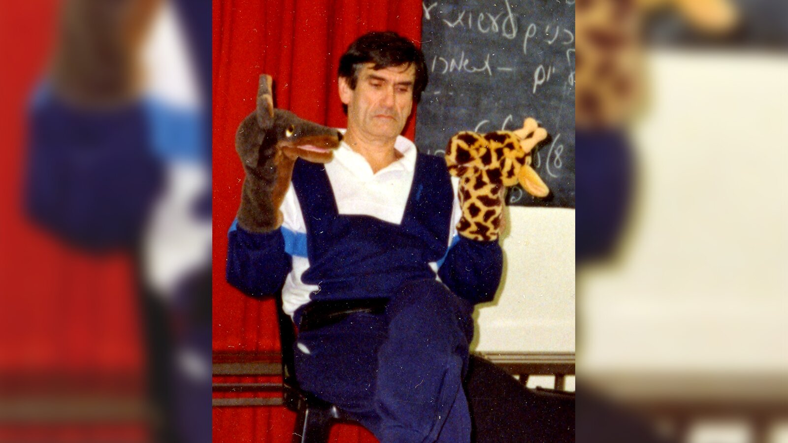 Ilustracja przedstawia aktora siedzącego na krześle. Na jego dłoniach widoczne pacynki wilka i żyrafy. 