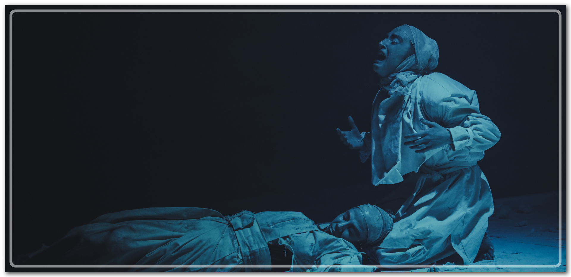 Zdjęcie przedstawia dwie kobiety wcielające się w role na scenie. Jedna z nich leży, druga siedzi blisko niej, na jej twarzy widać silne emocje.