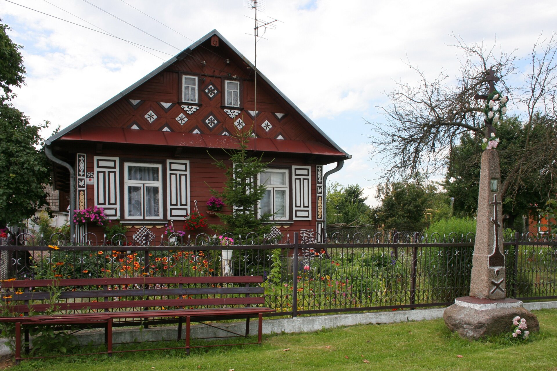 Zdjęcie przedstawia drewniany dom jednorodzinny wybudowany w ogrodzie. Dom, ze spadzistym dachem, ma ozdobne okiennice oraz rzeźbienia na fasadzie.   