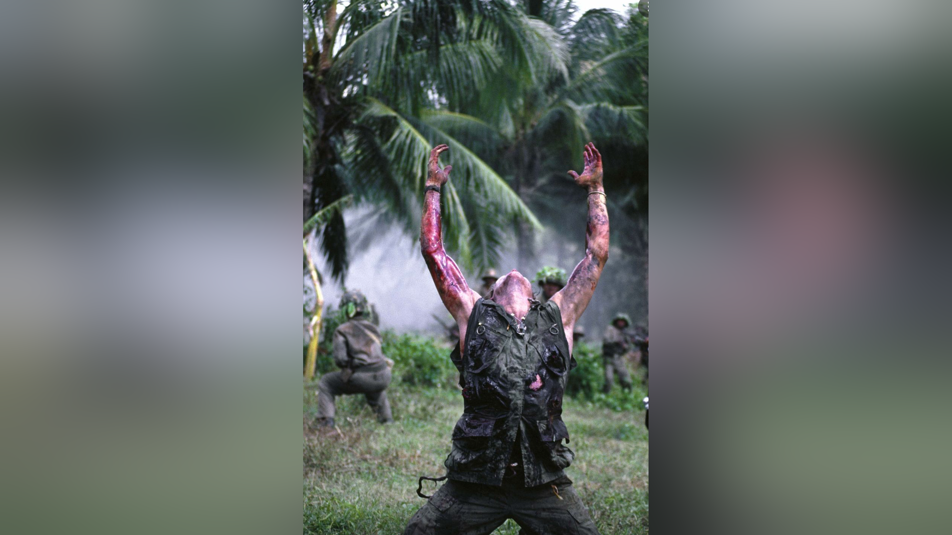 Zdjęcie przedstawia żołnierza, który klęczy, ma uniesione ręce i odchyloną do tyłu głowę. Na jego rękach jest krew. Jest kamizelka moro jest miejscami poszarpana. Za nim czają się żołnierze w maskujących strojach z bronią. W tle są plamy oraz unoszący się dym.
