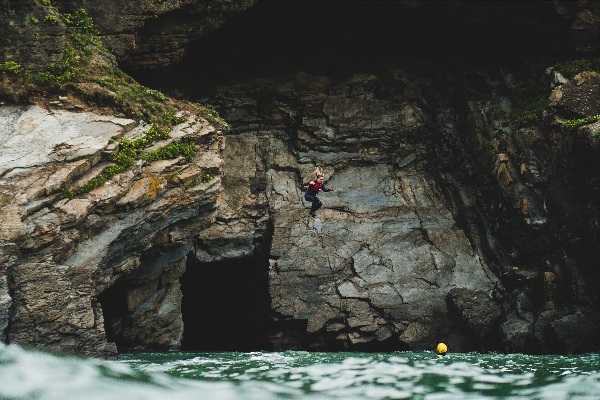 Zdjęcie przedstawia kobietę wykonującą skok ze skały do wody. Postać jest ubrana w piankę do pływania oraz kapok, ma rozłożone na boki ręce i ugięte kolana. W tle widoczna jest ściana skalna z półkami skalnymi i ciemnym otworem wlotowym jaskini. Na dole widoczna jest spieniona tafla wody. Po prawej stronie w wodzie widoczny jest żółty kask osoby, która prawdopodobnie asekuruje skok.  