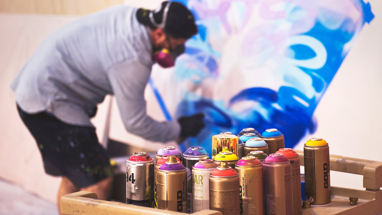 Zdjęcie przedstawia osobę z założoną maską na twarzy rysującą coś na ścianie. Widoczne są też pojemniki z farbami w sprayu. 