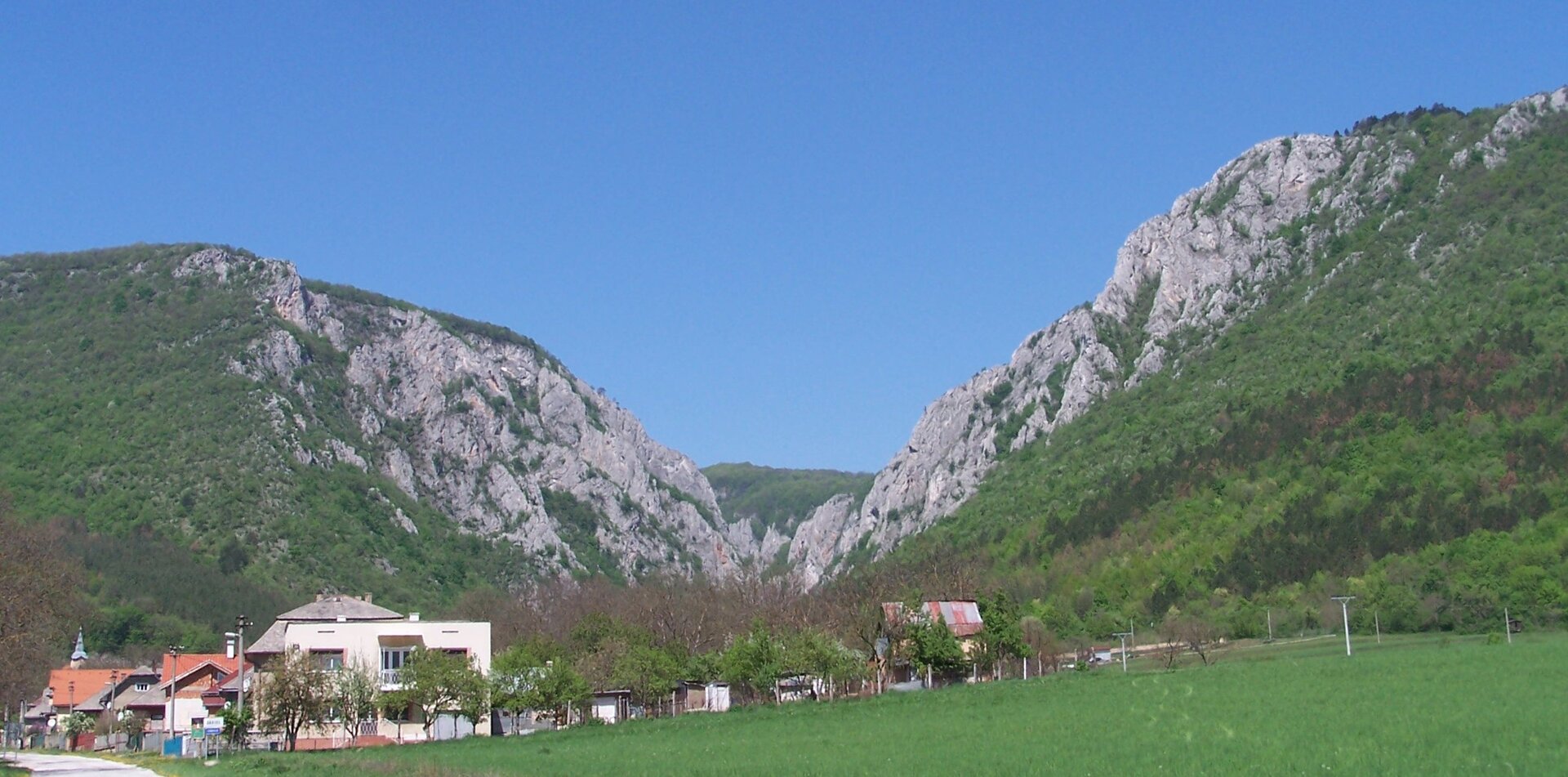 Na zdjęciu dolina. W tle dwa skaliste wzniesienia. Na dnie doliny znajdują się zabudowania mieszkalne wraz z dodatkową infrastrukturą. Dom na pierwszym planie otoczony jest ogródkiem i niskimi drzewkami. 