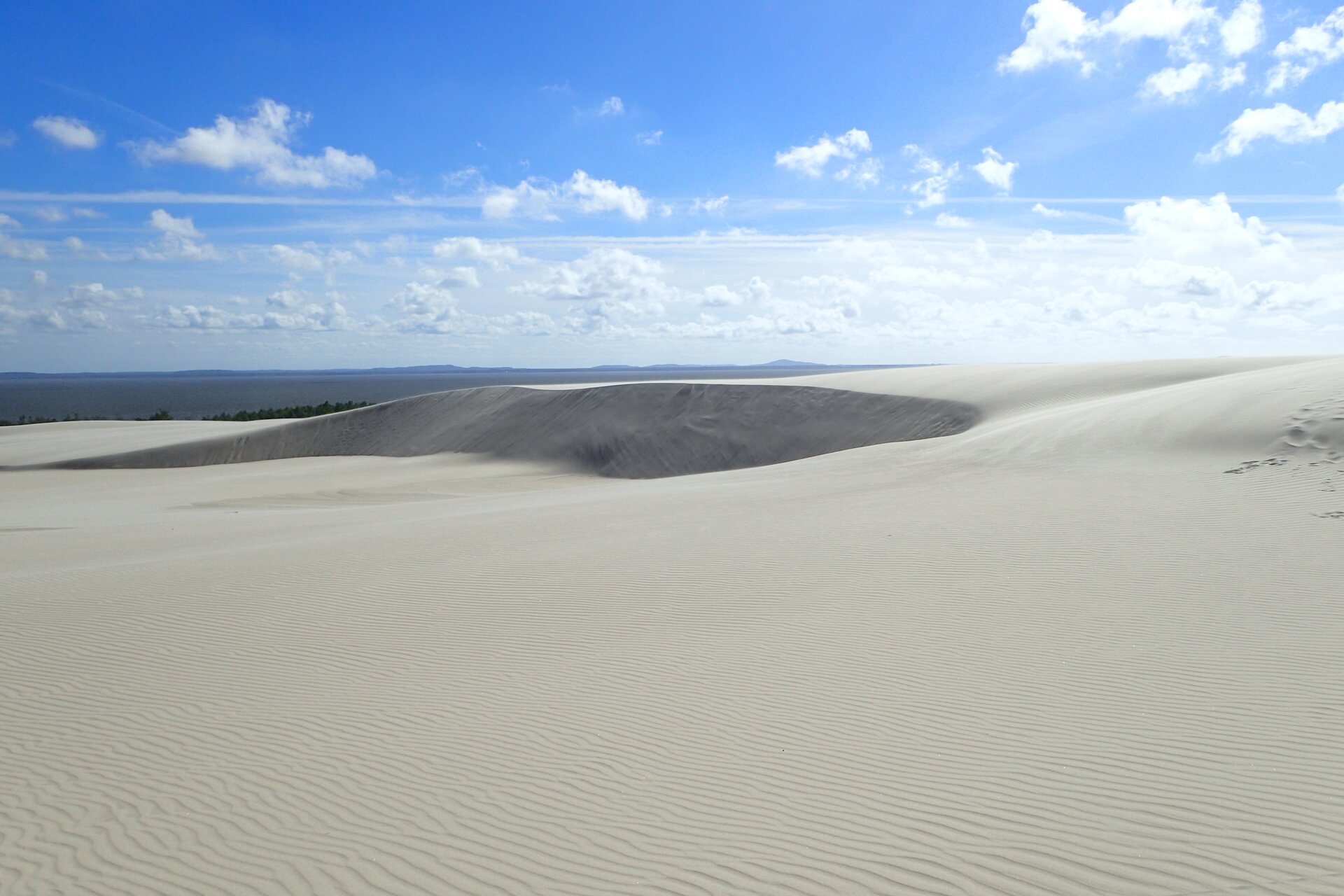 Zdjęcie przedstawia duży obszar pokryty jasnym piaskiem. Piasek tworzy wzniesienie - wydmę. W oddali widoczny jest zbiornik wodny. Na dalszym planie widoczne wzgórze. Nad wydmą niebieskie niebo, na którym są białe chmury. 