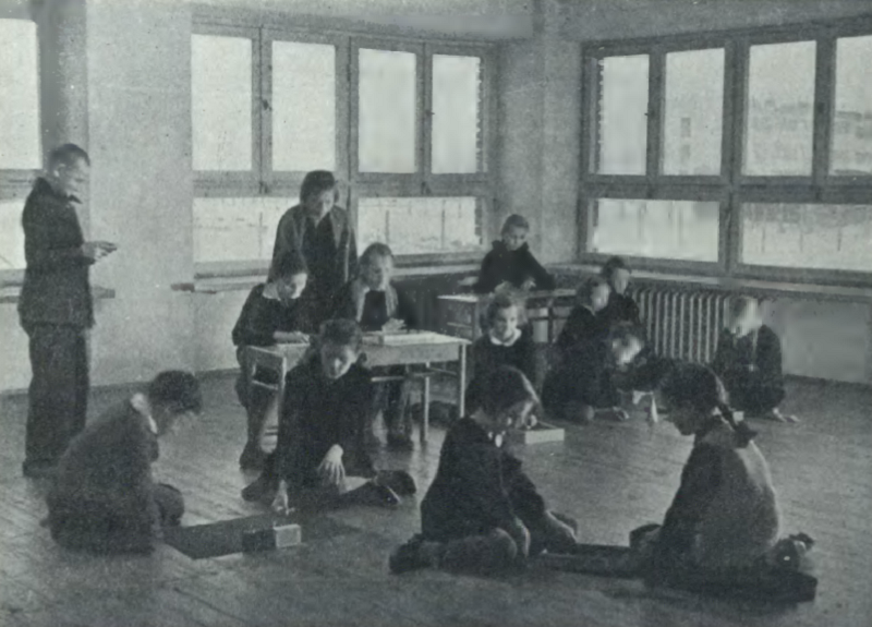 Zdjęcie przedstawia grupę dzieci i nauczycielkę w pomieszczeniu. Część dzieci siedzi na podłodze i gra w gry, część siedzi w ławkach i pisze w zeszycie. W tle stoi nauczycielka, pomaga dzieciom piszącym w zeszytach.