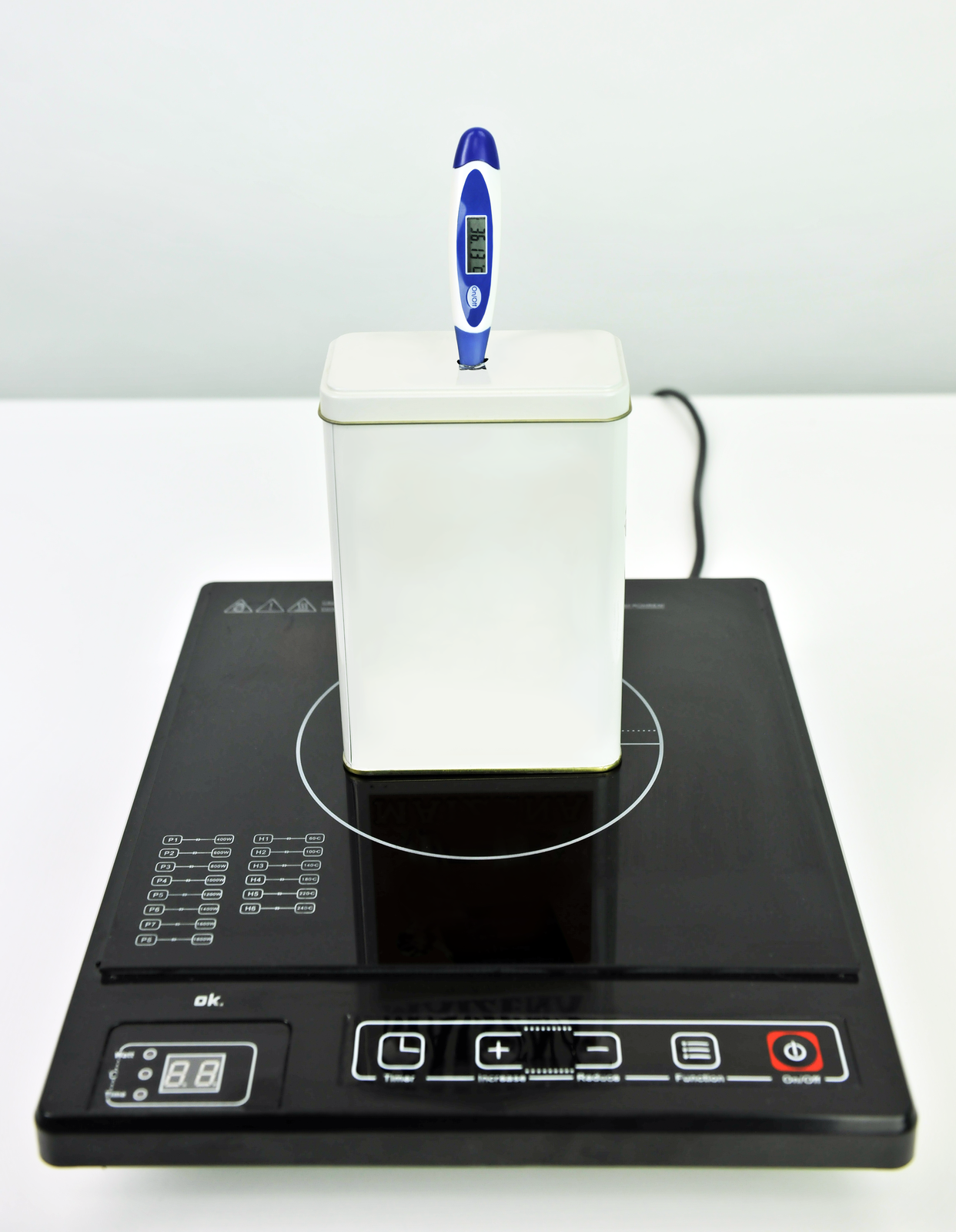 Fotografia przedstawia prostopadłościenne białe naczynie z pokrywą stojące na czarnej prostokątnej płytce elektrycznej. W pokrywie znajduje się termometr elektryczny. Tło jasne.