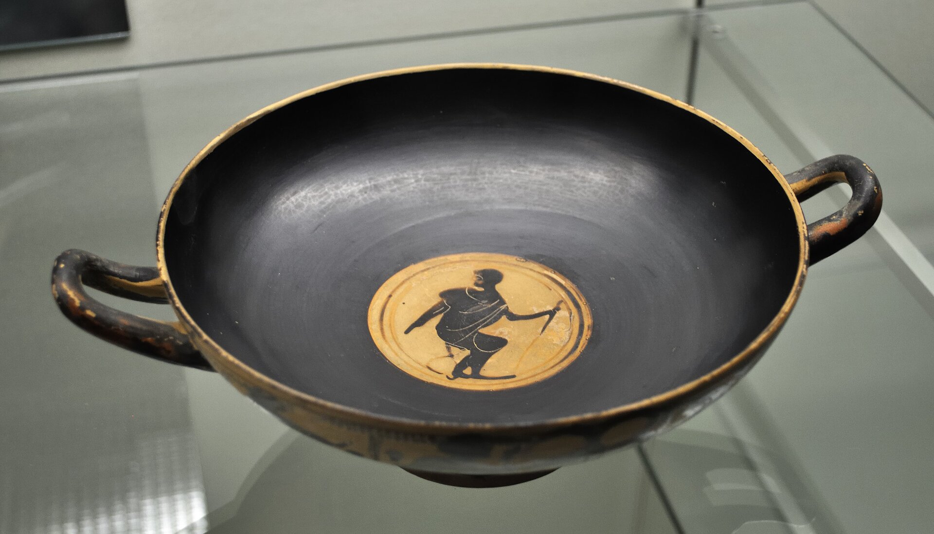 Fotografia przedstawia naczynie antyczne w stylu czarnofigurowym. Ma ono kształt wazy. Na dnie naczynia na żółtym tle przedstawiono czarną postać męską ze sztyletem w ręku.