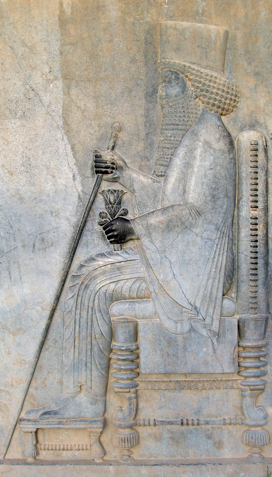 Zdjęcie przedstawia relief z wizerunkiem Dariusza I, władcy perskiego, który brał udział w wojnie z Grekami. Dariusz został ukazany jako mężczyzna w podeszłym wieku, o czym może świadczyć długi zarost. Mężczyzna siedzi na tronie, na jego głowie spoczywa korona, odziany jest w długie szaty. W jednej dłoni trzyma długą laskę, w drugiej zaś puchar kształtem przypominający tulipan. Przedstawiony został z lewego profilu.
