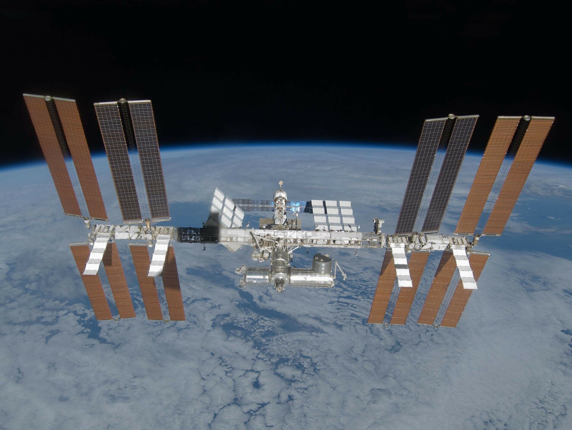 Na zdjęciu jest międzynarodowa stacja kosmiczna. To ogromna konstrukcja, której korpus złożony jest z wielu cylindrycznych modułów i długiej szyny, do której przyczepione są skrzydła z baterii słonecznych. Po prawej i po lewej stronie stacji są po dwie pary pionowych skrzydeł z bateriami słonecznymi powyżej szyny i po dwie pary poniżej szyny. W tle stacji kosmicznej widoczny jest fragment kuli ziemskiej, pokrytej chmurami.
