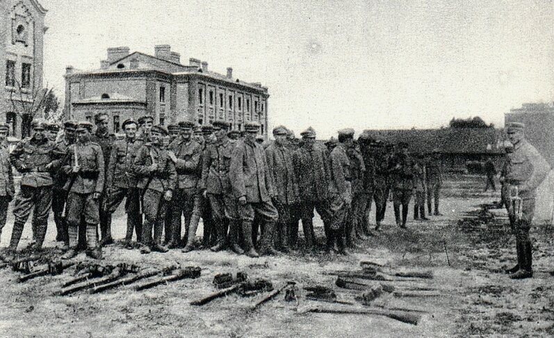 Zdjęcie przedstawia grupę żołnierzy stojących na placu. Przed nimi leży broń. W tle stoją dwa budynki. Przed żołnierzami stoi mundurowy z wąsem. 