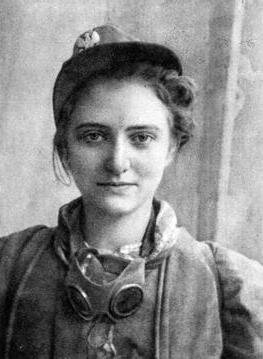 Kliknij, aby powiększyć. Zdjęcie przedstawia młodą dziewczynę. Na głowie ma czapkę z orzełkiem. Nosi marynarkę. Na szyki ma chustę i okulary. Jest uśmiechnięta.