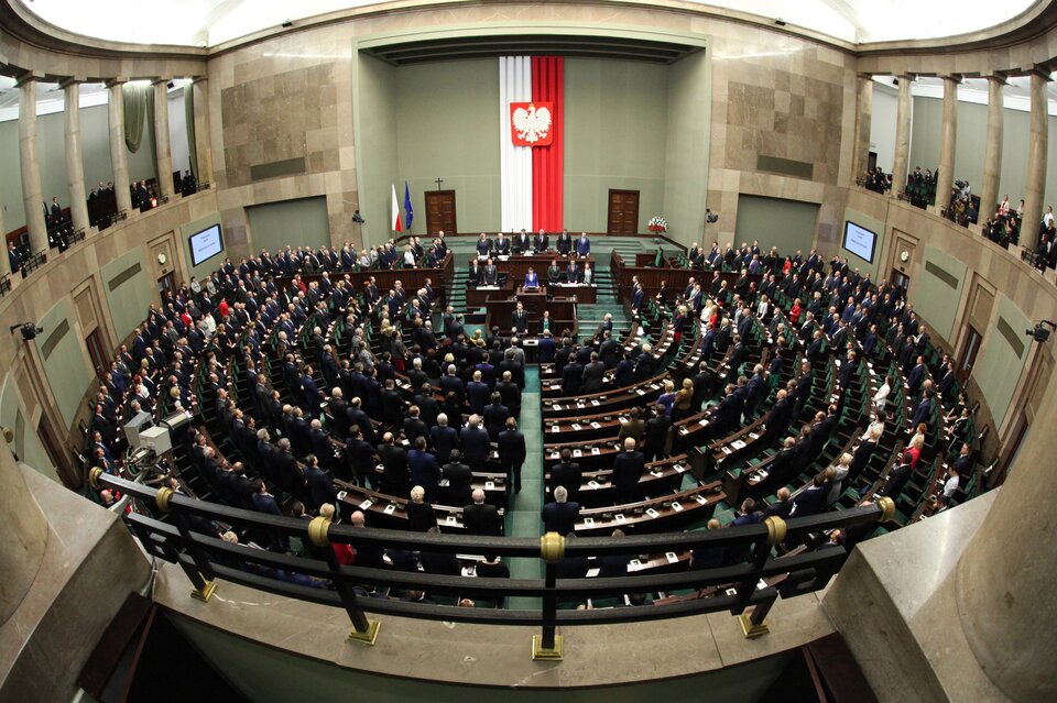 Zdjęcie przedstawia salę obrad polskiego Sejmu. Sala jest wypełniona posłami, którzy stoją. Sala ma kształt półkola - tu znajdują się ławy poselskie. Na końcu sali, na podwyższeniu zasiada Marszałek Sejmu, a po jego prawej stronie Rada Ministrów. Przed stołem Marszałka stoi mównica dla posłów.