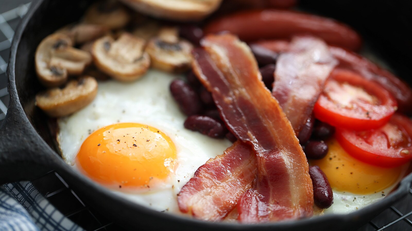 Na zdjęciu widoczna jest patelnia, na której widoczne są dwa jajka sadzone, spieczone paski bekonu, czerwona fasola, talarki pieczarek i pomidory pokrojone w plastry.