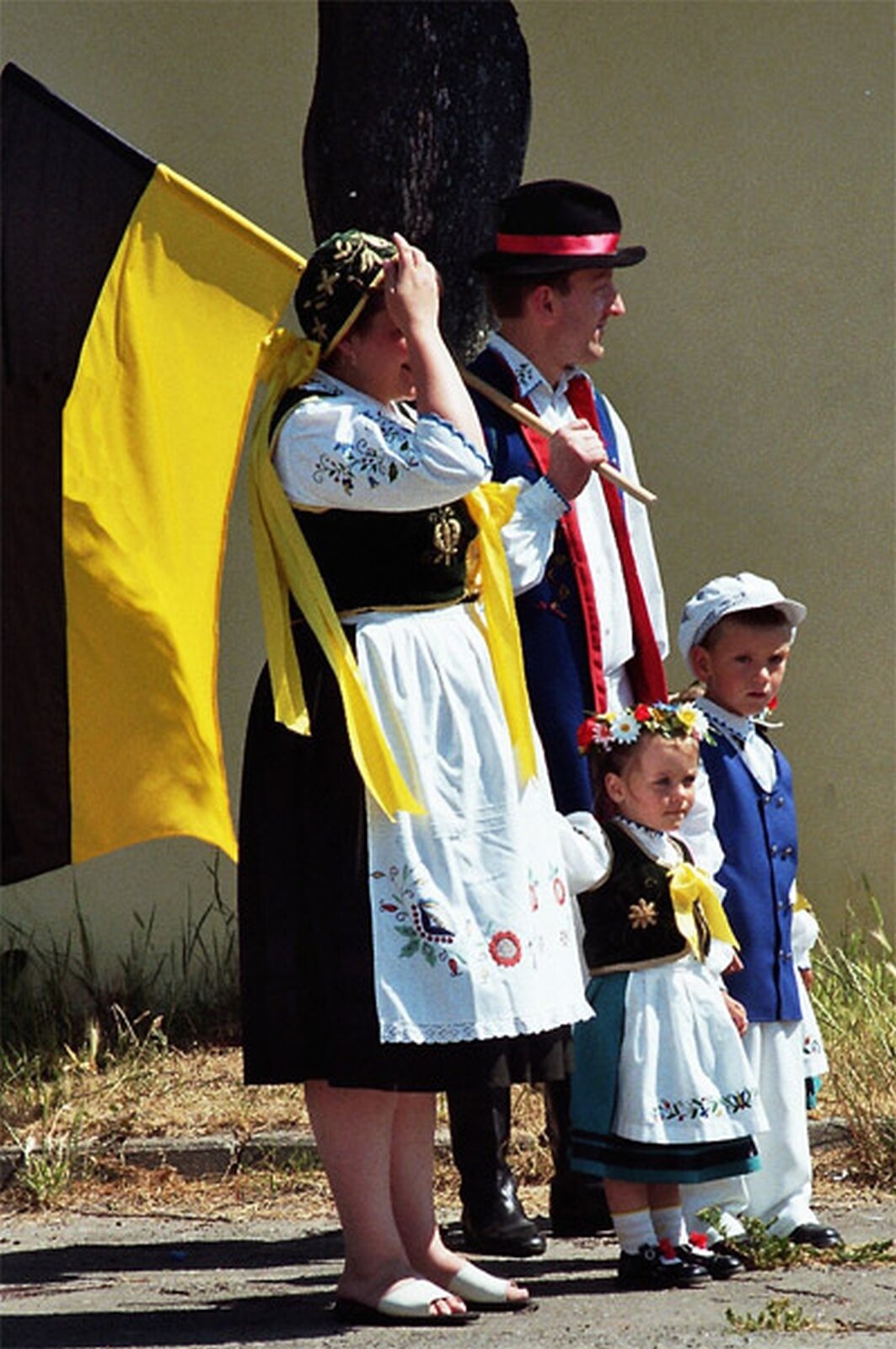 Zdjęcie przedstawia rodzinę kaszubską. Kobieta, mężczyzna, mała dziewczynka i nieco wyższy od niej chłopiec. Noszą tradycyjne stroje. Mężczyzna opiera na ramieniu maszt dużej czarno‑żółtej flagi.