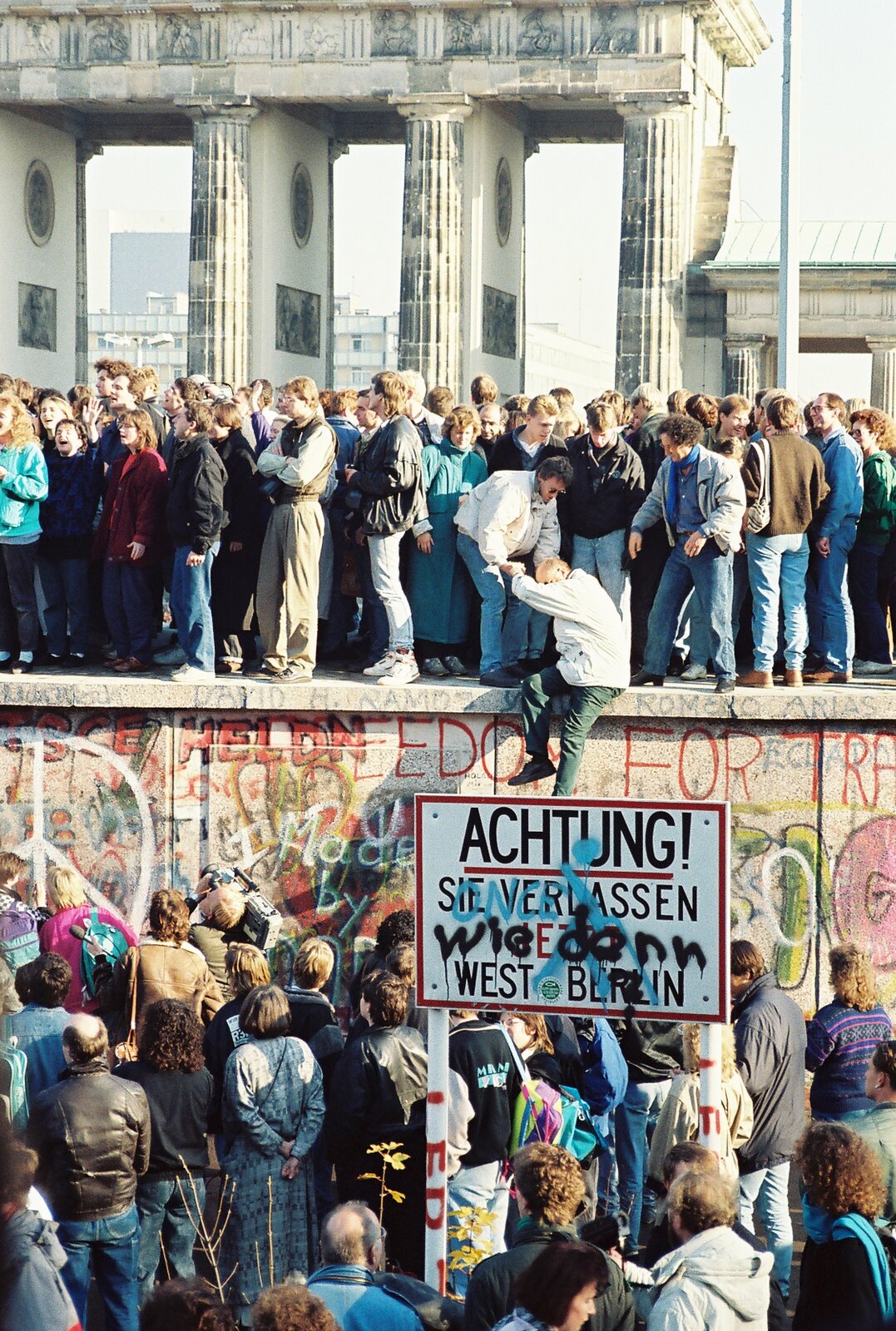 Zdjęcie przedstawia ludzi tłumnie zgromadzonych na szczycie oraz u podstawy muru berlińskiego w okolicach Bramy Brandenburskiej. Wśród zgromadzonych są kobiety, mężczyźni i dzieci. Jedna z osób stojących na murze pomaga wspiąć się na niego starszemu mężczyźnie. Ściana muru pokryta jest kolorowymi napisami i ściennymi grafikami. Przed nim ustawiona jest tablica ostrzegawcza z napisem w języku niemieckim, na której dopisane są czarnym sprayem dodatkowe słowa.