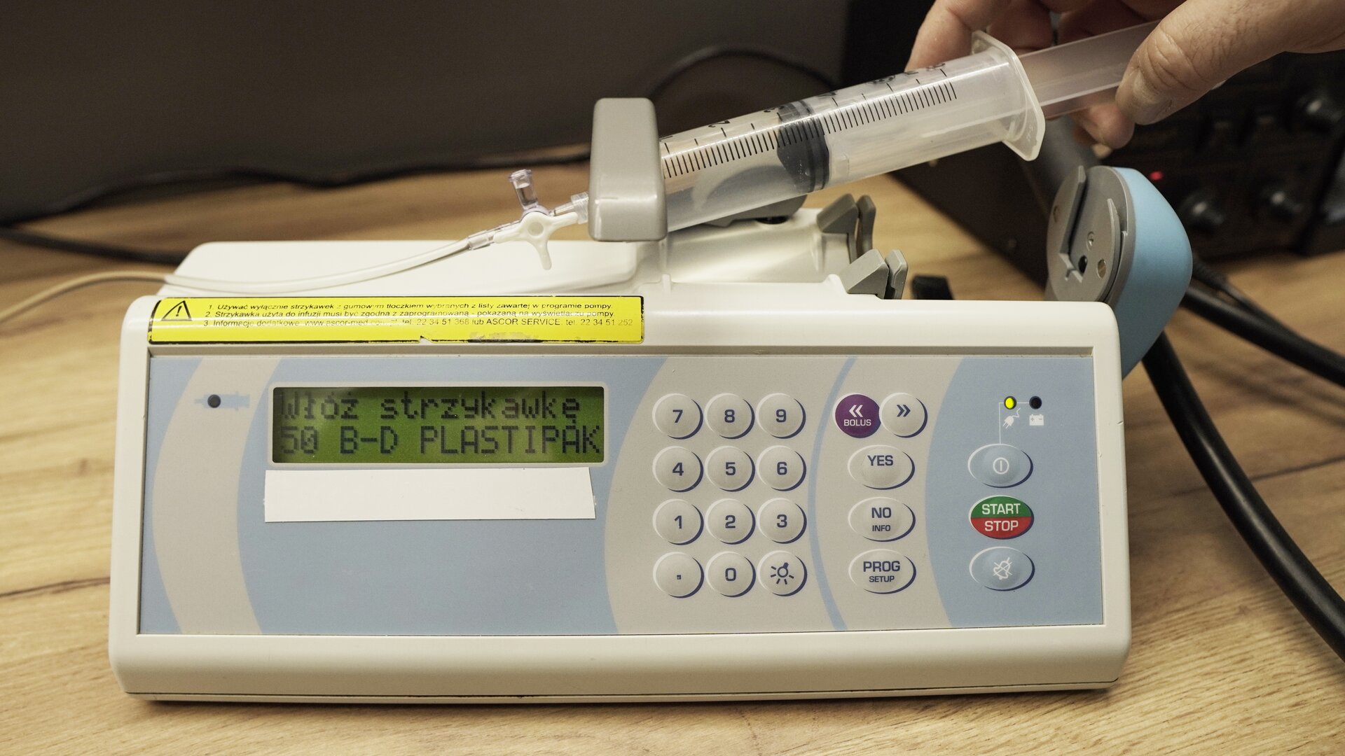 Na zdjęciu technik wkłada strzykawkę do ramienia pompy infuzyjnej. Na ekranie aparatu wyświetlana jest informacja: "Włóż strzykawką 50 B‑D PLASTIPAK".