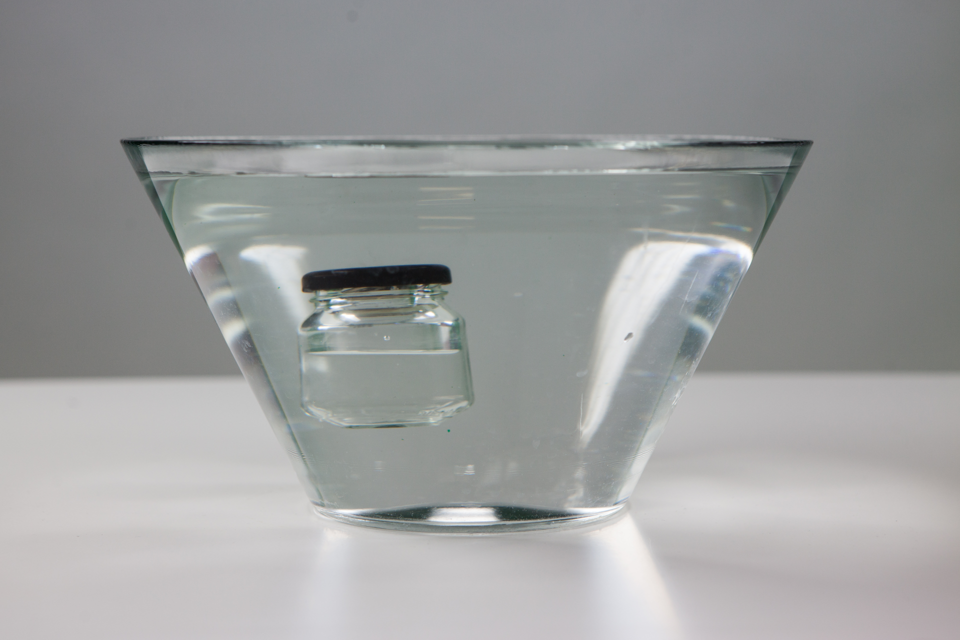 Zdjęcie przedstawia mały zakręcony słoiczek napełniony wodą i odrobiną powietrza, unoszący się w dużym pojemniku z wodą powyżej dna, ale poniżej powierzchni – ujęcie z boku. Słoiczek znajduje się pośrodku większego naczynia.