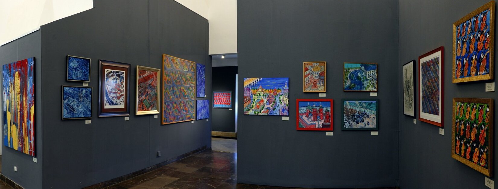 Fotografia przedstawia wystawę obrazów, które znajdują się na grafitowej ścianie. Po prawej stronie znajduje się dziewięć obrazów. Po lewej stronie, na ściennie wisi siedem obrazów. Wszystkie działa różnią się od siebie wielkością. 